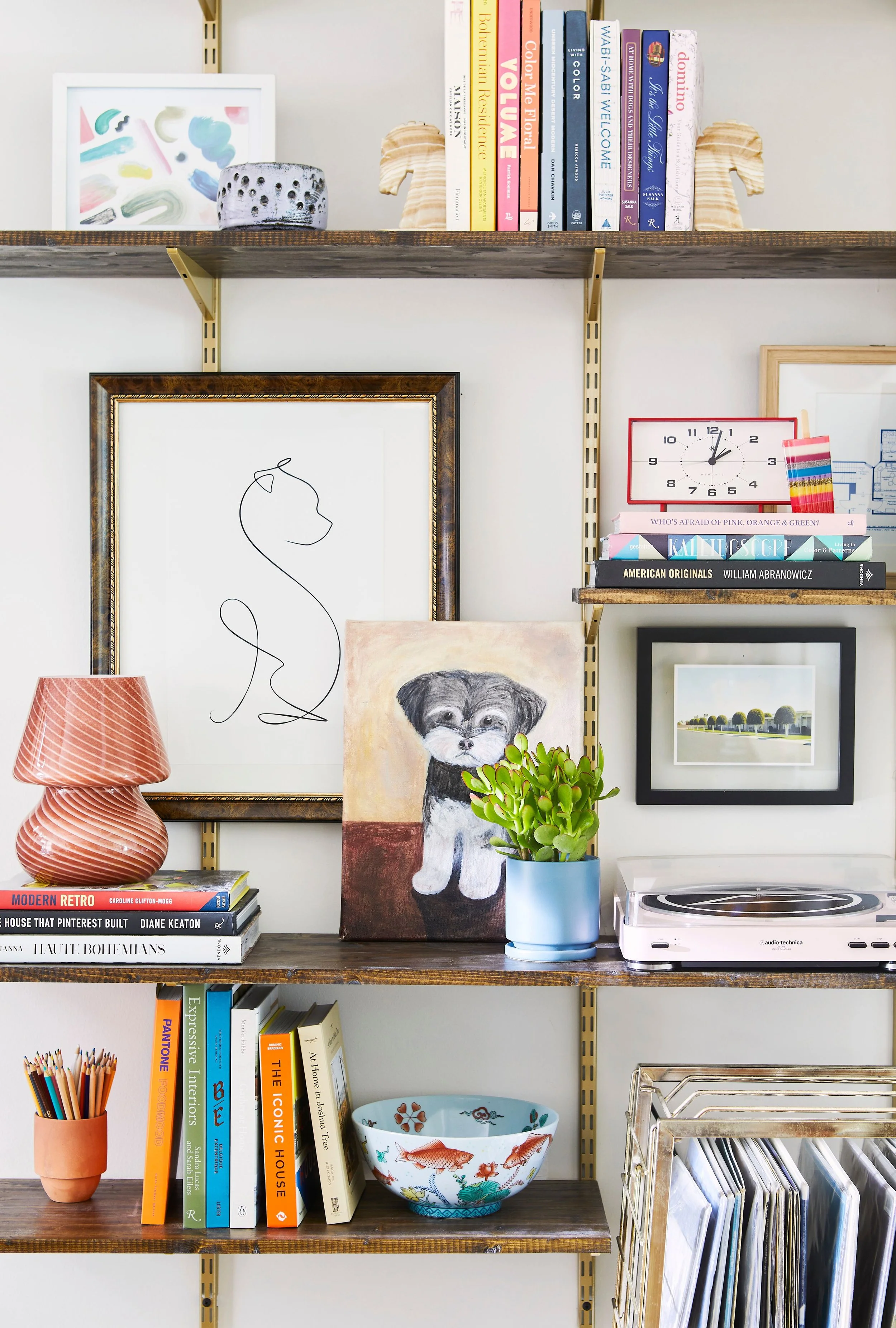 Colorful wall-mounted shelving styled with books, playful art, a dog painting, small lamp, plant, and record player for a bright eclectic look.