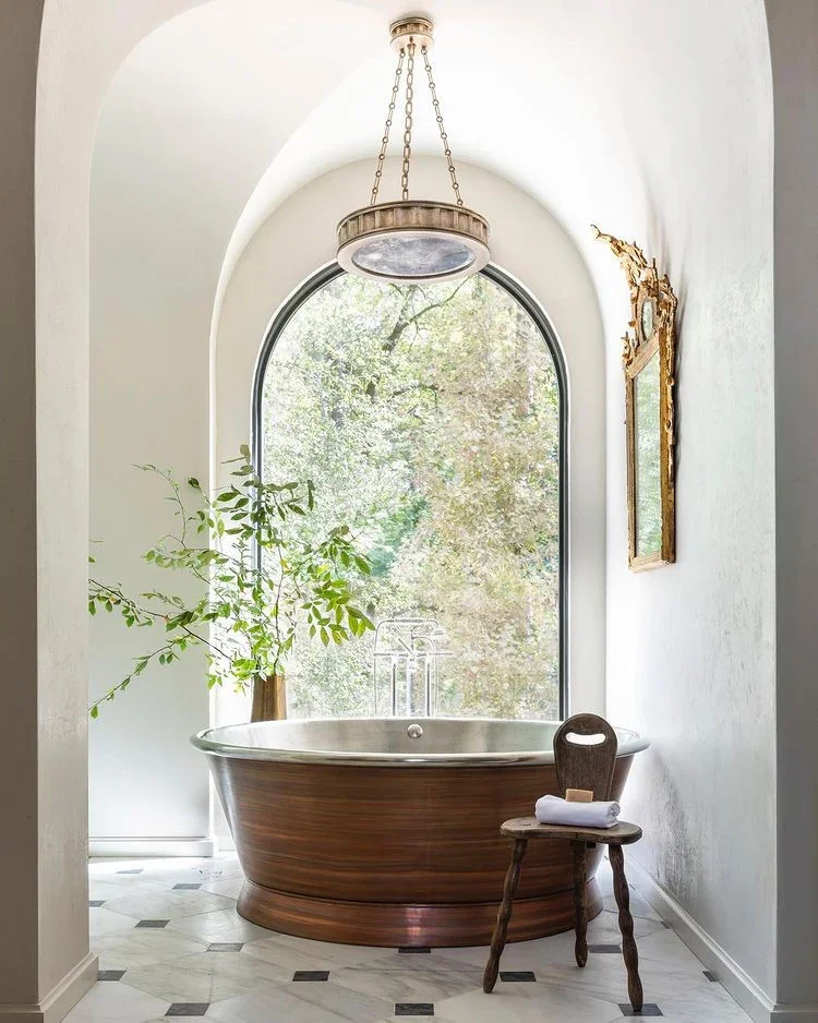A well illuminated European-inspired bathroom with a large arched window behind the soaking tub, plaster walls, marble floors, and natural light make this bath shine with light.