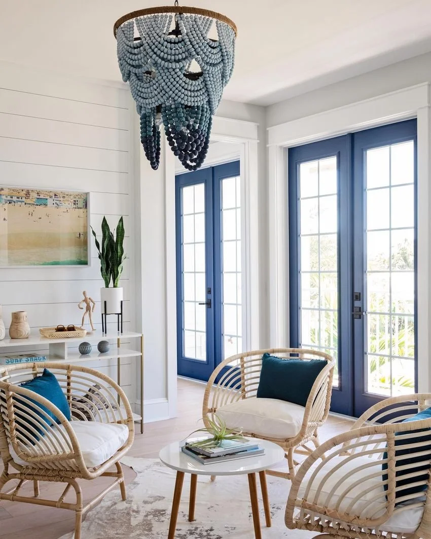 Coastal sitting room design with pale rattan lounge chairs, cushy teal pillows, round white table, and blue beaded chandelier in front of blue French doors.