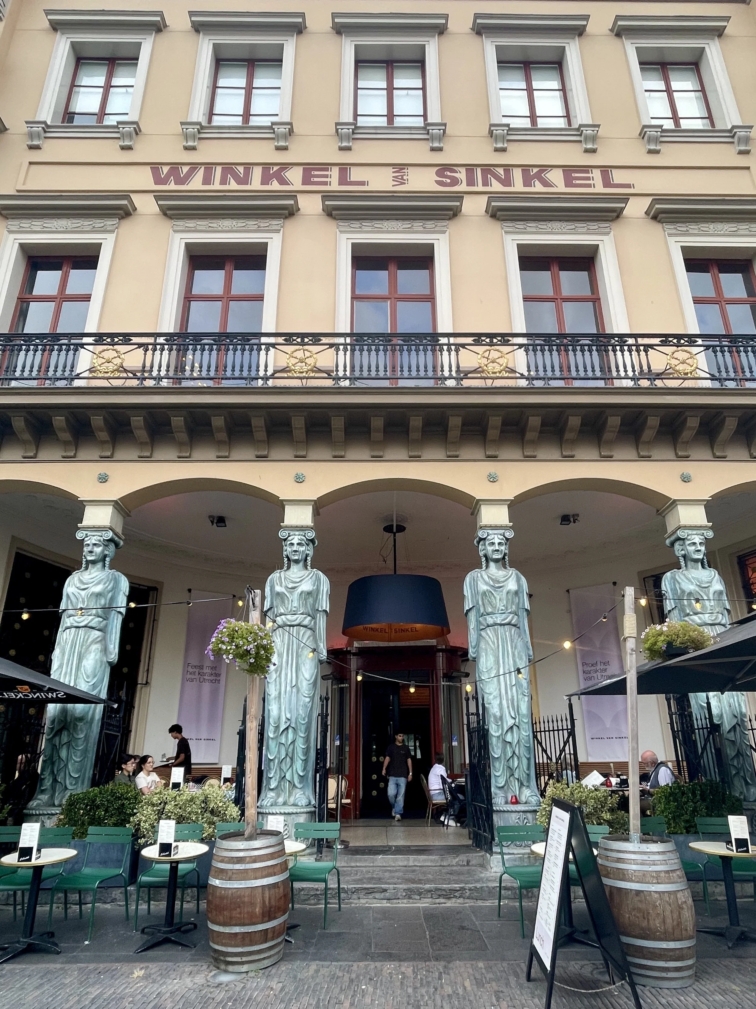 The Neoclassical building of Winkel van Sinkel in Utrecht has yellow paint with red windows and lettering, a fancy wrought iron terraced second level and green sidewalk chairs.