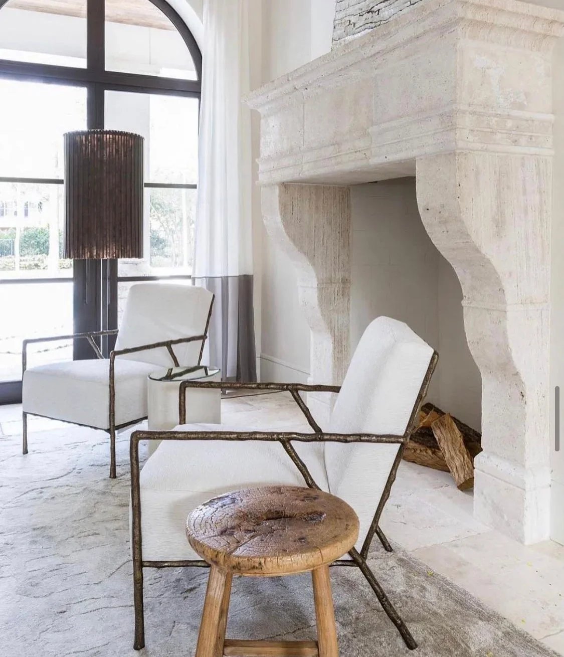 Daylight in a European-inspired sitting room with arched window, stone fireplace, linen chairs, and filtered natural light.