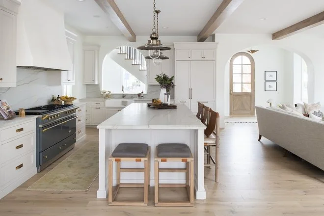 European-inspired kitchen with white cabinetry, marble island, wood beams, arched openings, and natural light borrowed from adjoining rooms.