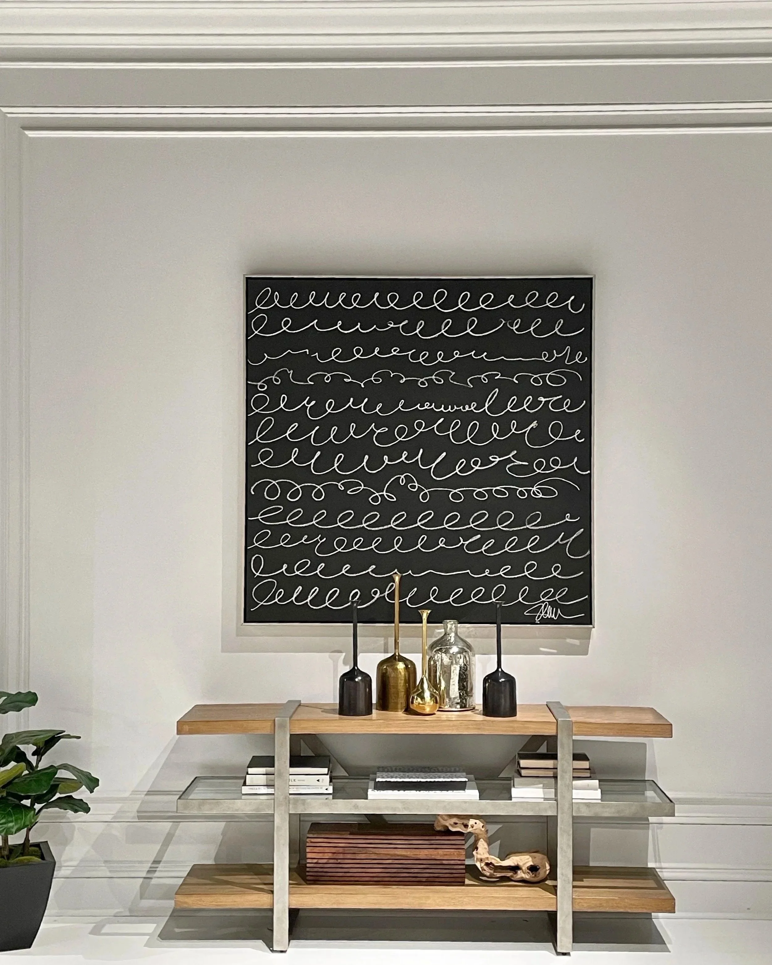 A luxury living space features a wood and steel console table with three shelves decorated with accessories. A dramatic black canvas painting with white script hangs above. Paneled walls reflect classic design. Natural potted plants are on left.