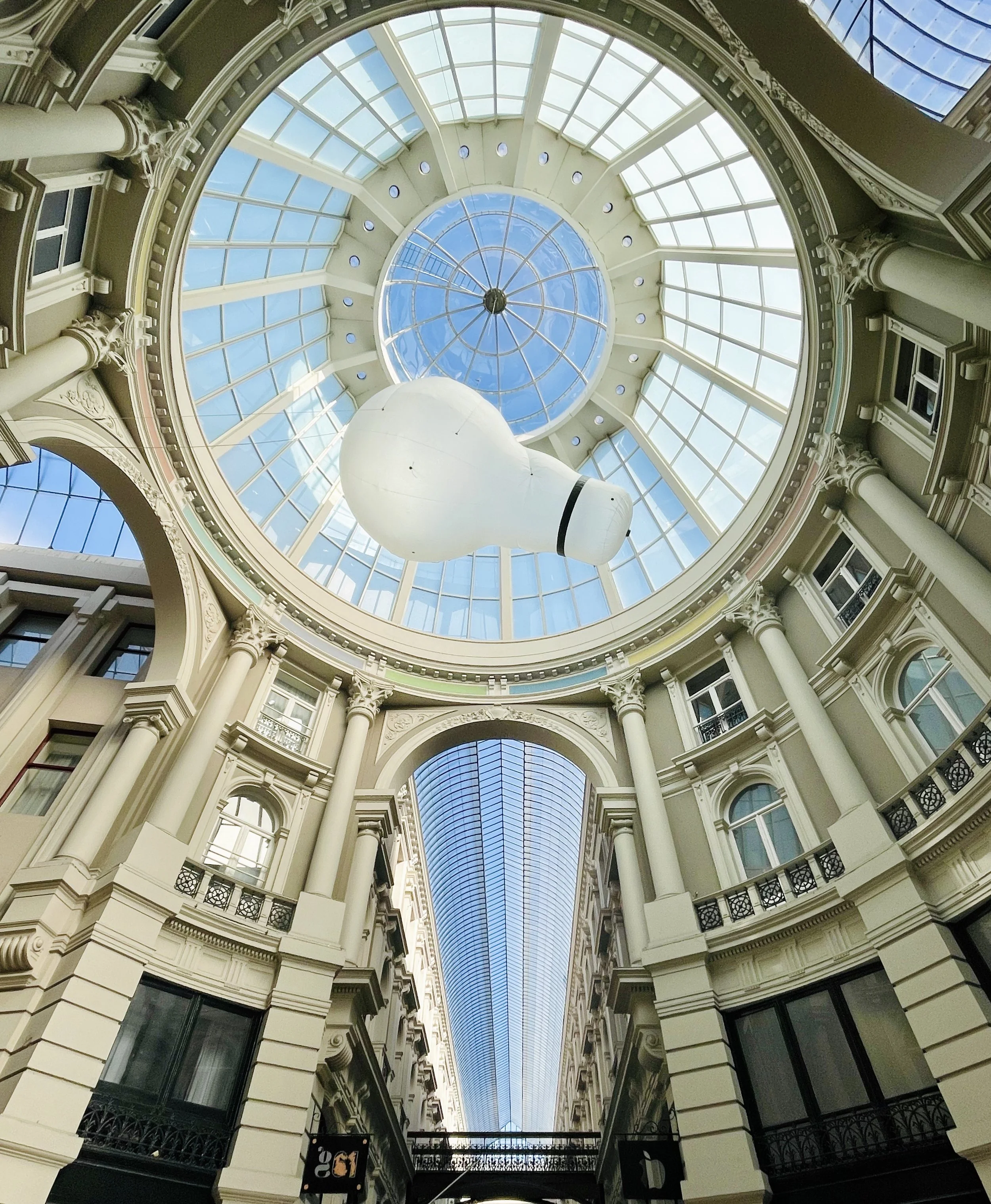 Interior view of De Passage in The Hague showing a glass dome ceiling with a large white lightbulb sculpture and classical columns.