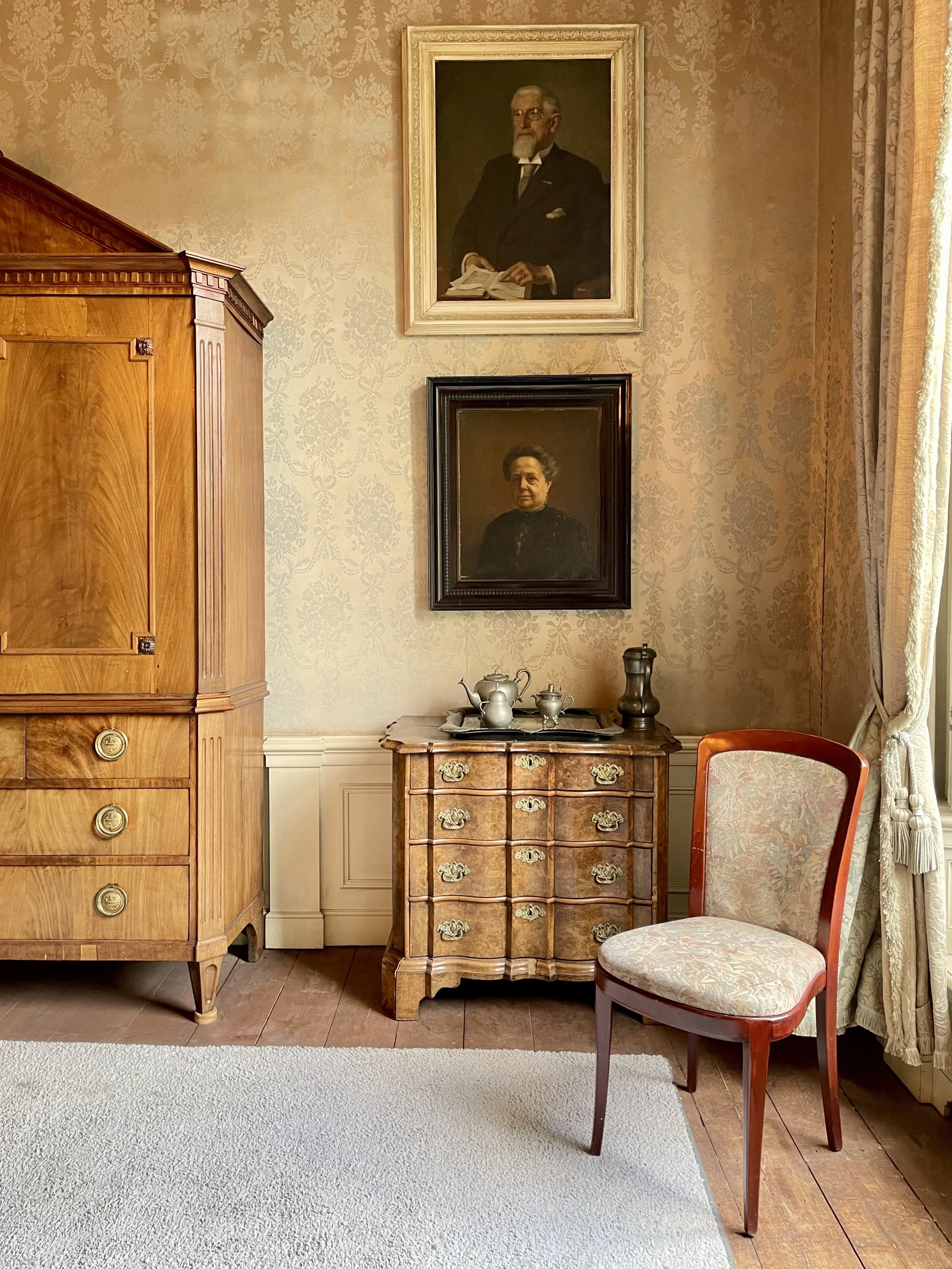 Corner of historic room with antique wood furniture, framed portraits, and upholstered chair