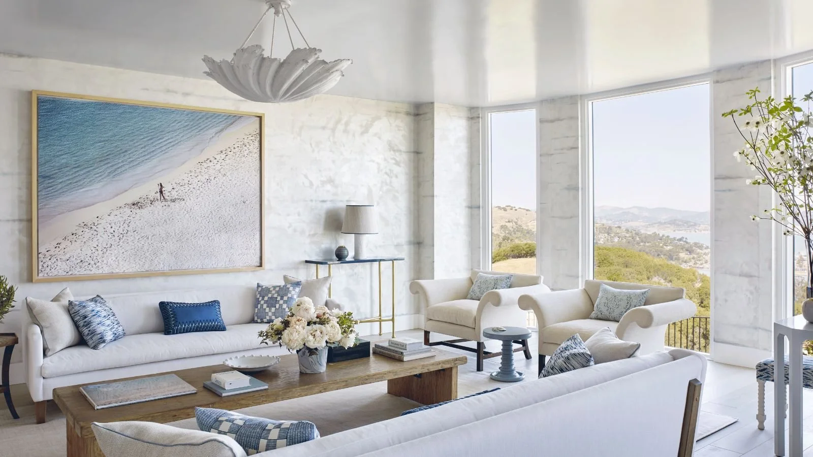 Palm Beach light coastal living room with white sofas, white plaster chandelier, blue accents, modern photography of the beach, and water view.