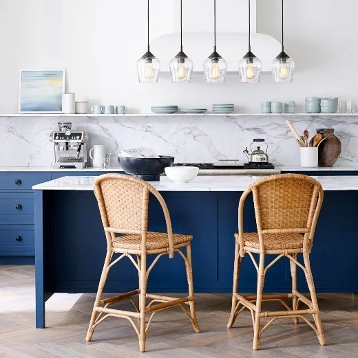A bright coastal kitchen features a deep navy island with a white marble countertop, two woven rattan barstools, a marble backsplash, open shelving with pale blue dishes, and a row of clear glass pendant lights above.
