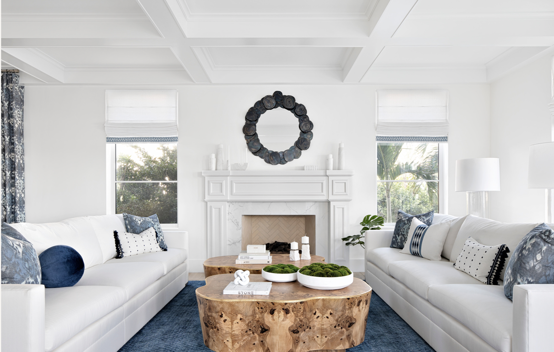 Luxury Palm Beach living room with white sofas, organic live-edge wood coffee tables, blue rug and pillows, and round sculptural mirror above a white fireplace.
