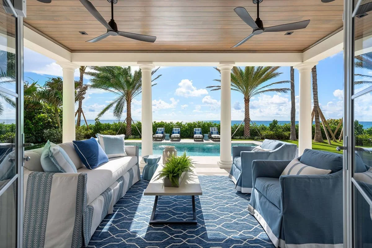 Luxury Palm Beach home coastal outdoor living room with ocean views, pool, white columns, blue furniture, and wood ceiling on the patio.