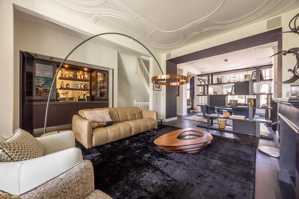 Contemporary Dutch living room with built-in bar, curved floor lamp, black rug, and modern shelving beneath ornate plaster ceilings.
