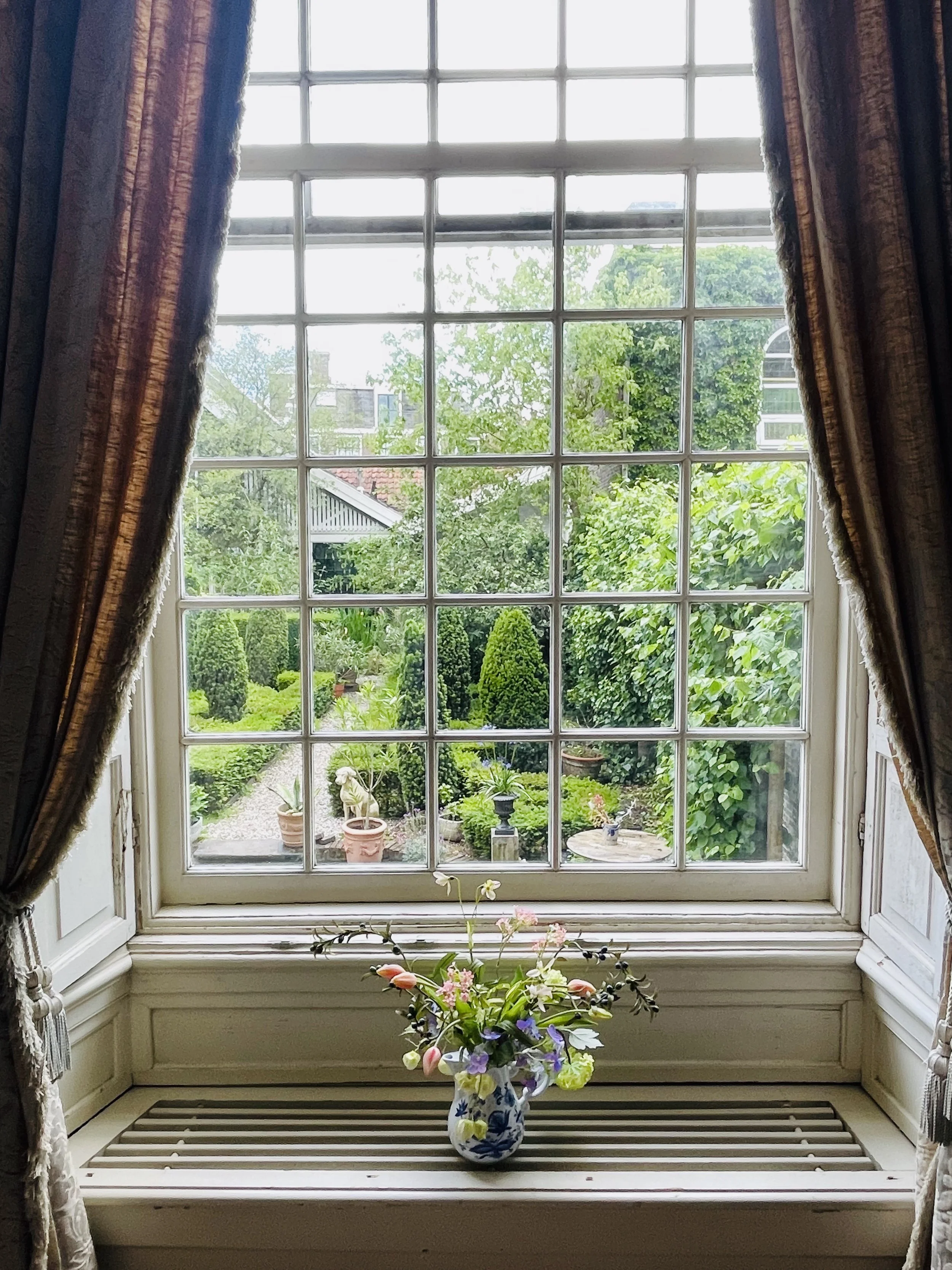 View through a divided-light window to a formal green garden with potted plants and clipped hedges.