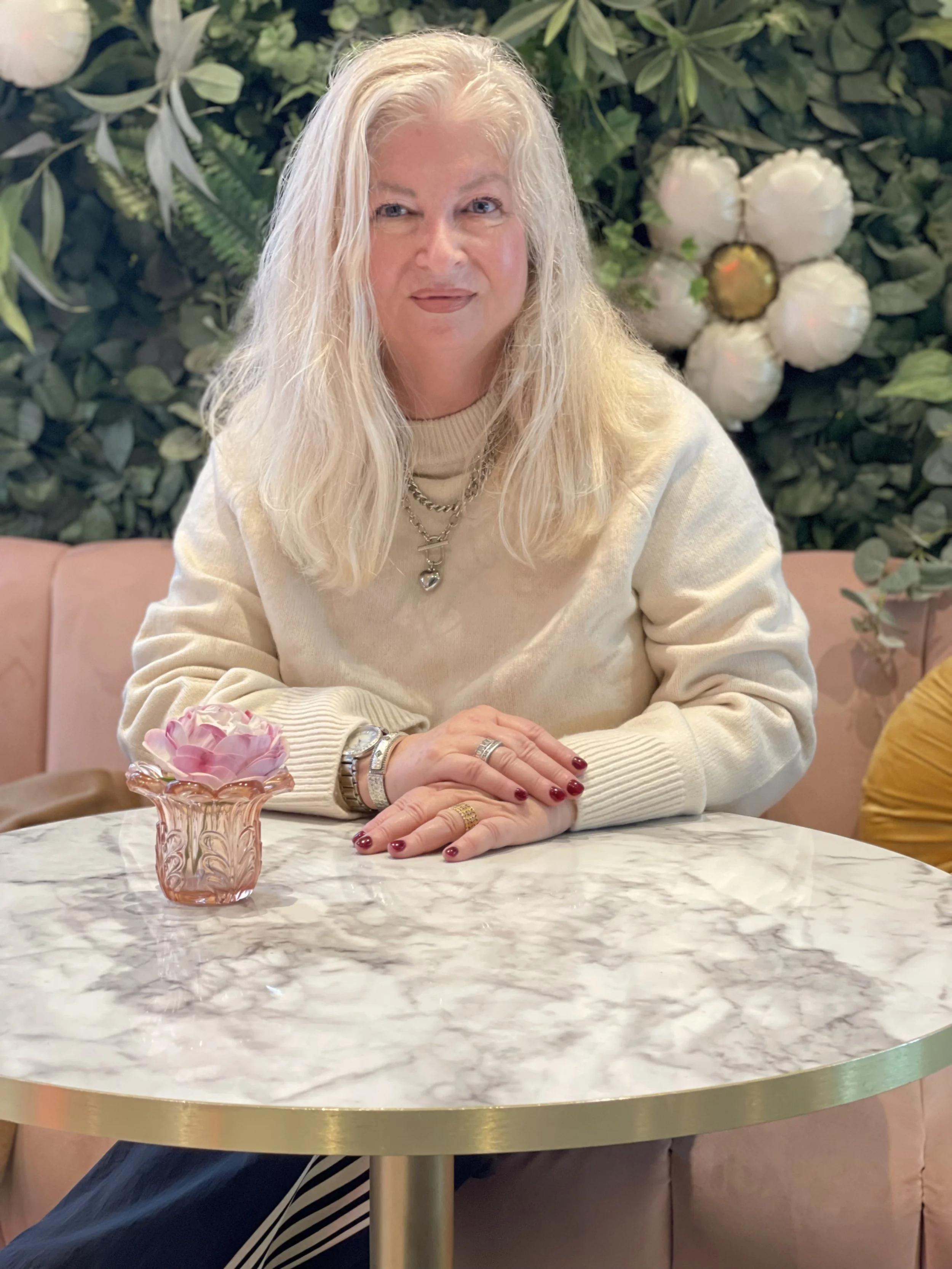 Woman seated at a marble café table in front of a lush green wall, smiling softly at the camera.