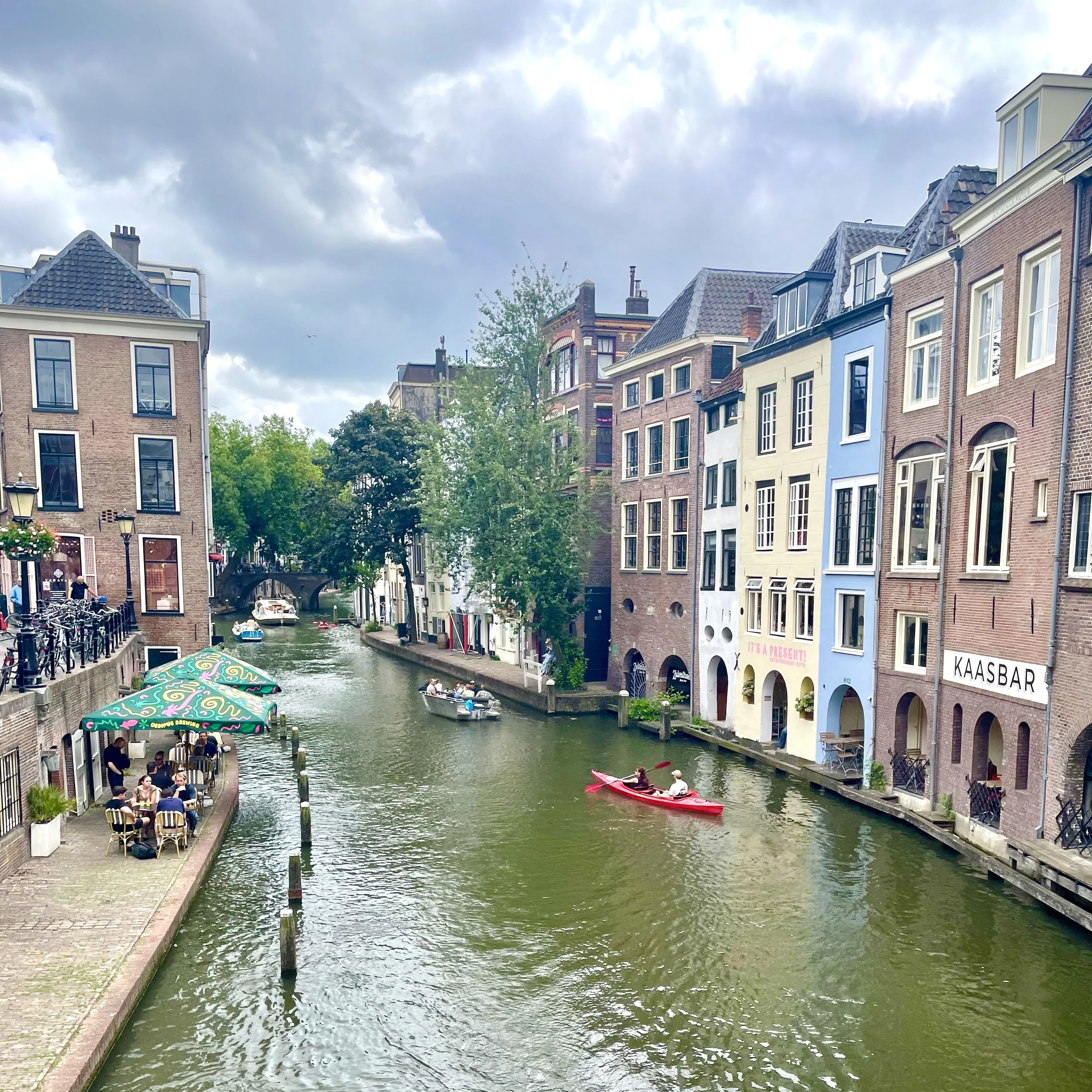 A beautiful Utrecht canal below street level shows some pastel painted and brick houses, some which house commerce, as people paddle in canoes and boat down the  canal.