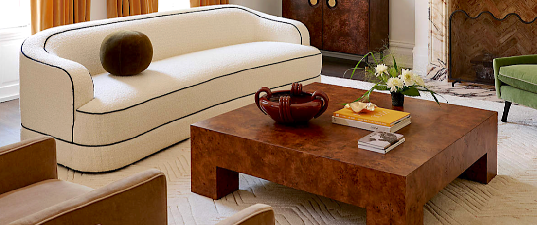 Palm Beach living room of luxury interior design shows a comfortable seating area with white modern sofa, trimmed in black with olive round pillow, on a neutral textured rug with burl wood square coffee table layered with flowers, books, more.