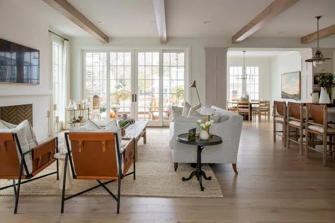 Light-filled open-concept living and dining room with French doors, wood beams, neutral furniture, and leather chairs