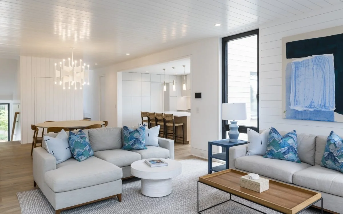 Modern coastal living room with wood accented walls, white modern chandelier, blue pillows, table and art, and open kitchen.
