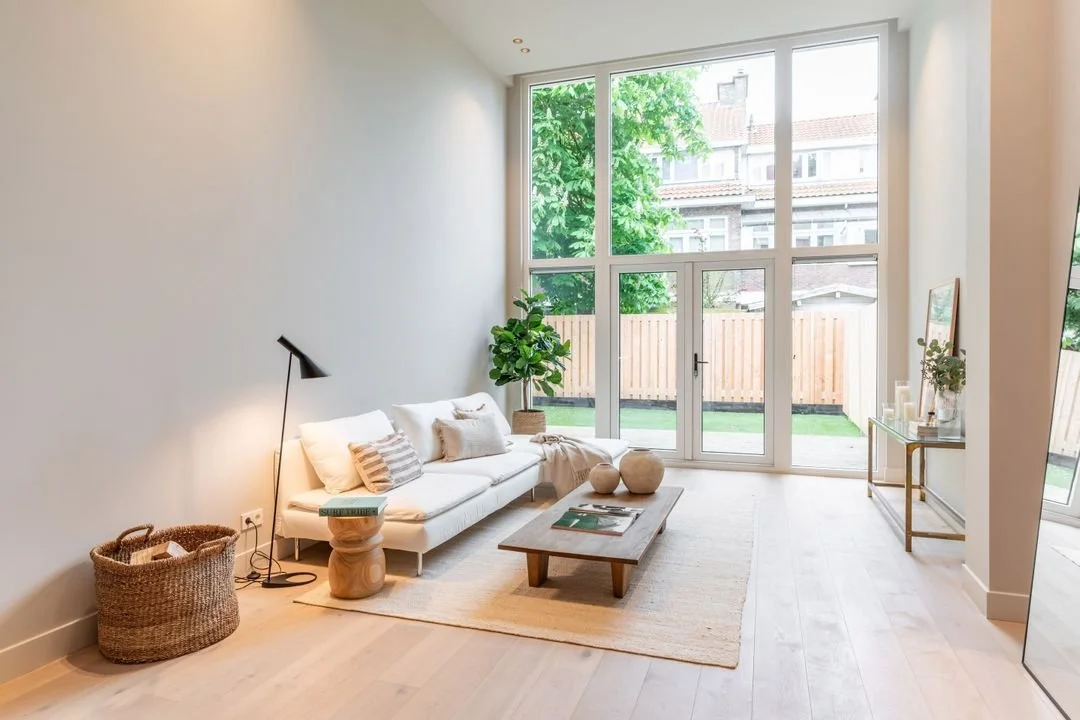 Bright minimalist living room with tall windows, white sofa, neutral rug, and wood coffee table opening to a small garden.