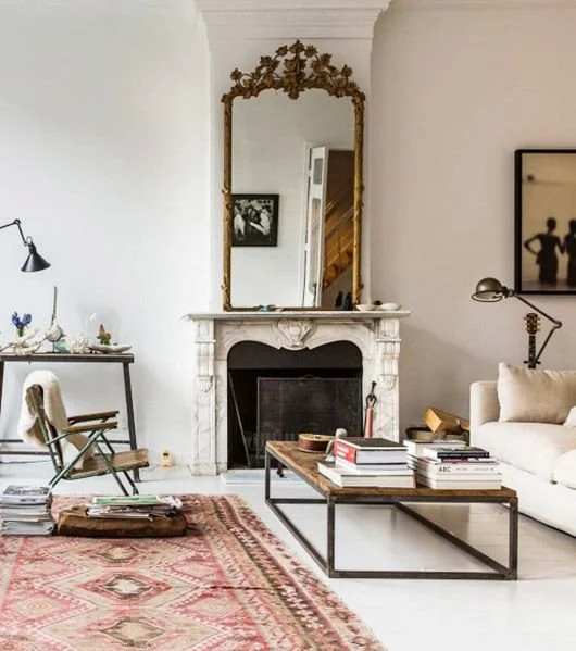 A Palm Beach coastal vintage-style living room with an ornate gold mirror above a marble fireplace, a neutral sofa, industrial-frame coffee table covered with books,