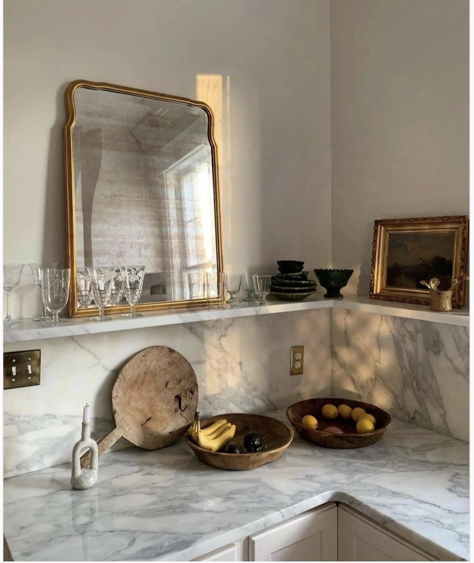Palm Beach marble countertop kitchen corner with open shelf, vintage gold-framed mirror, glassware, and wooden bowls of fruit.
