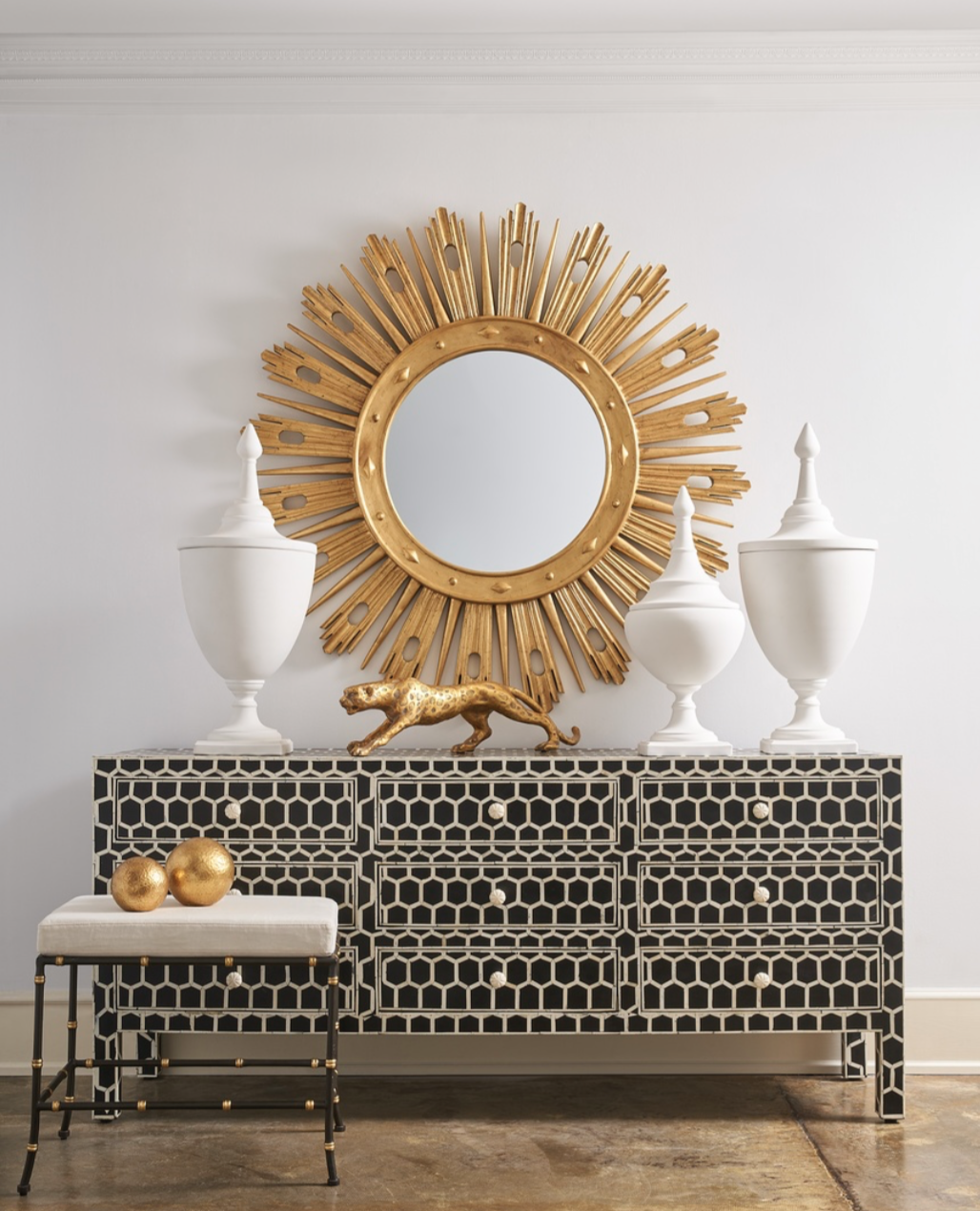 This black chest has bone inlay. A French vintage style sunburst gold mirror sets the entrance foyer scene for a warm welcome, and the chest is a perfect display top for white urns and a sleek golden panther. A small bench is perched in front.