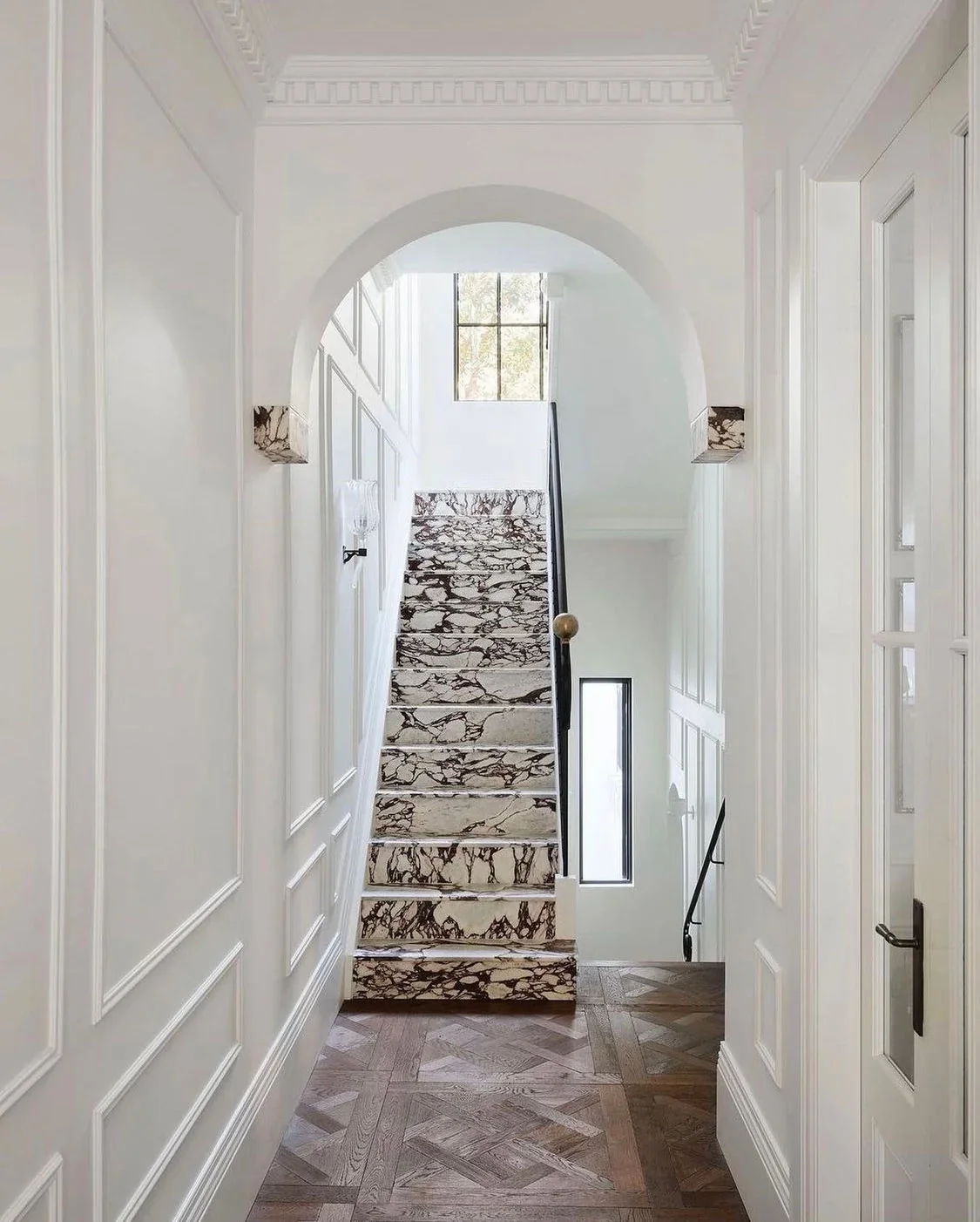 Elegant European stairway with marble steps, white millwork, natural light, and warm wood flooring.