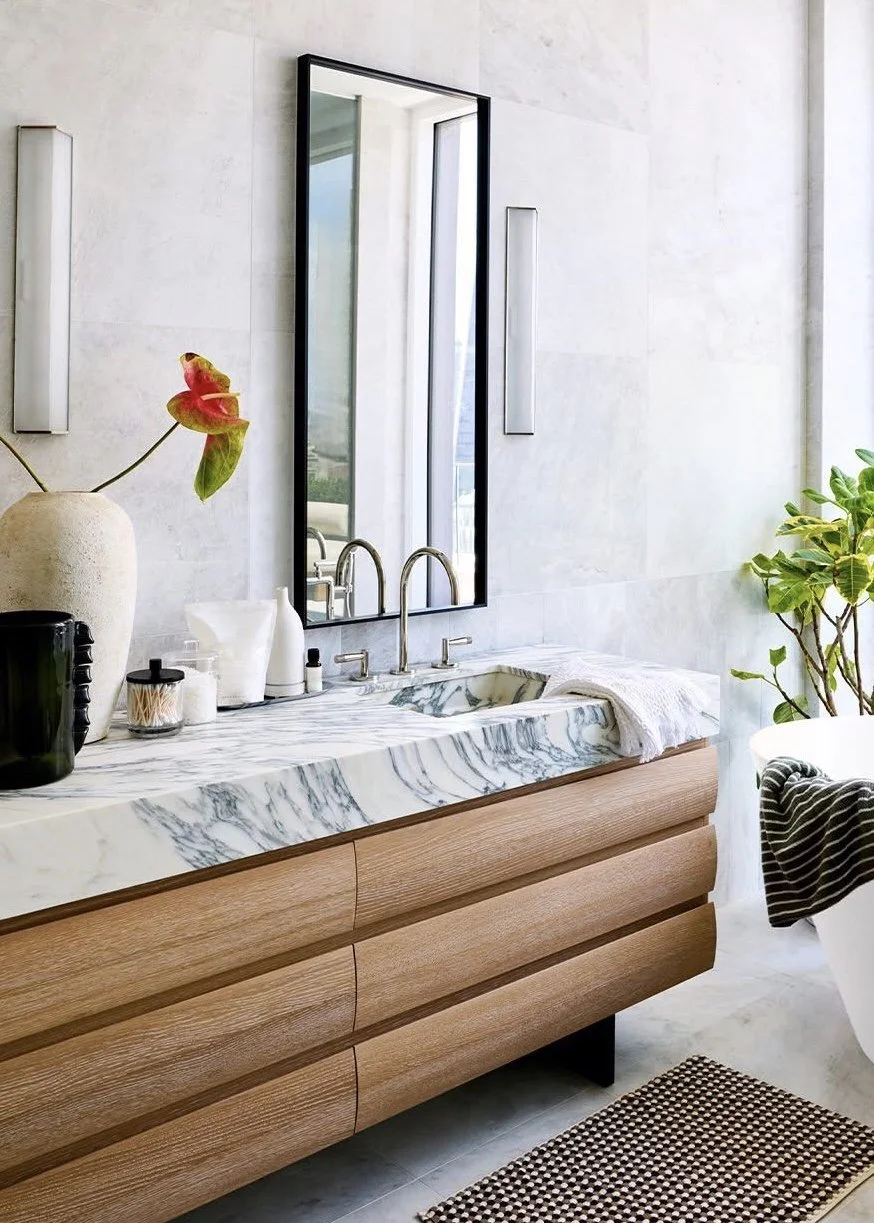 A Palm Beach bathroom has a rounded oak drawer vanity with striking white marble with black veins, and a tall black rimmed mirror flanked by sconces.