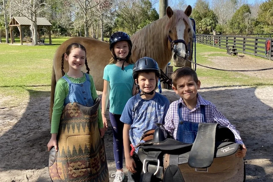 Four children in riding helmets holding saddles and pads beside a horse at Hidden Palms Ranch in Sanford