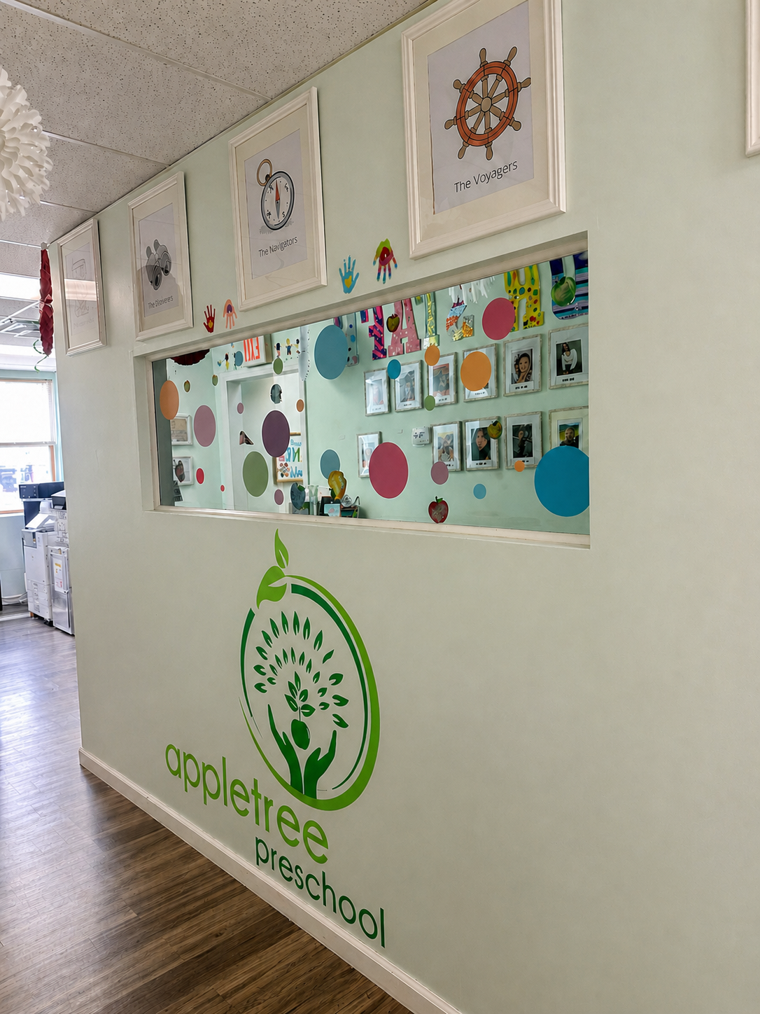 Interior wall of a preschool with colorful decorations, framed signs labeled "The Navigators," "The Voyageers," and a clock. Below the window, there is a large green logo and text reading "appletree preschool." The window has colored dots and cutouts, and in the background, a reception area with equipment and shelves.