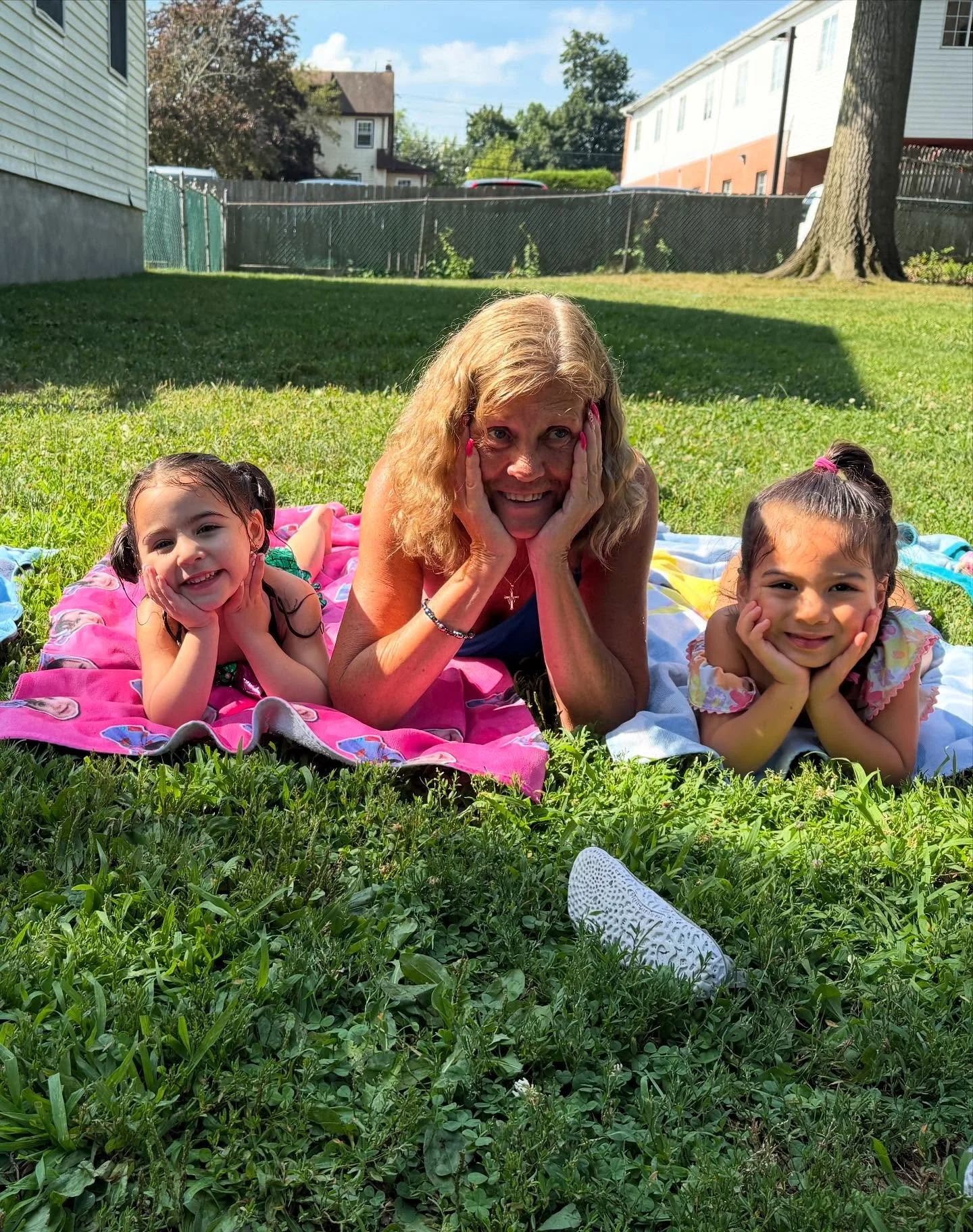 A woman with two young girls lying on colorful towels on a grassy yard, smiling and resting their chins on their hands in a sunny backyard with trees, houses, and a fence in the background.