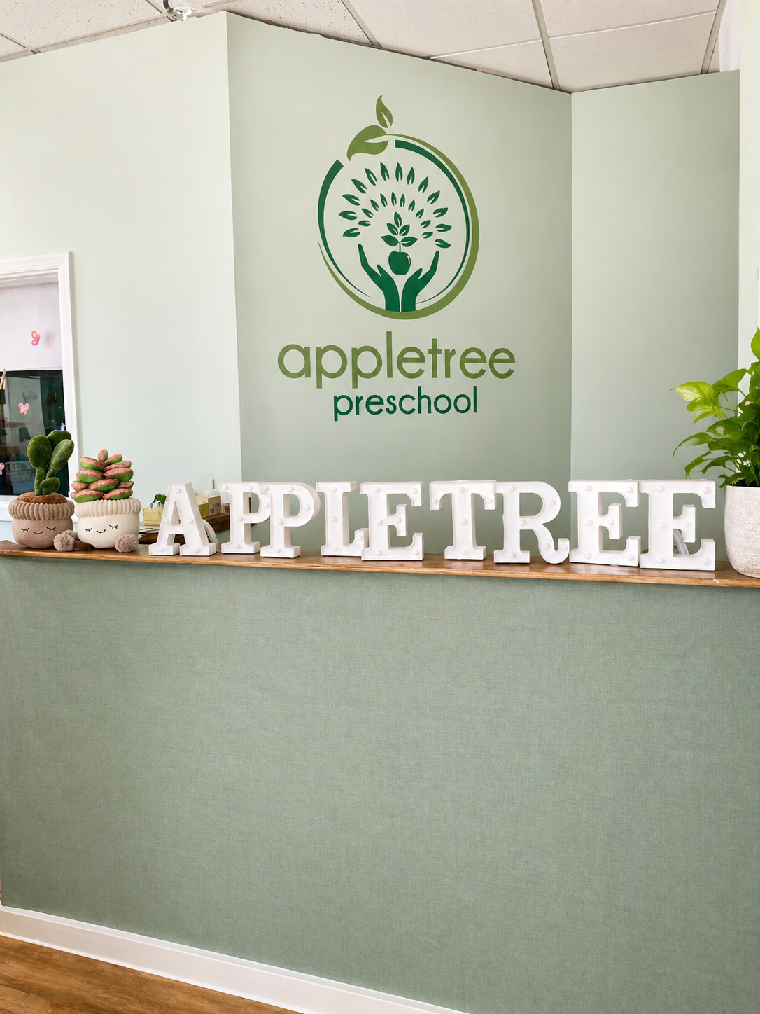 Reception desk with decorative plush plant pots and large white letter signage spelling 'APPLE TREE' in front of a green wall with a logo for 'appletree preschool' featuring a stylized tree and hands.