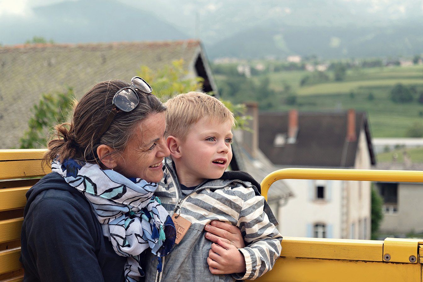 A woman and a young boy sitting on a yellow bench outdoors, looking at something with interest, against a rural landscape background.