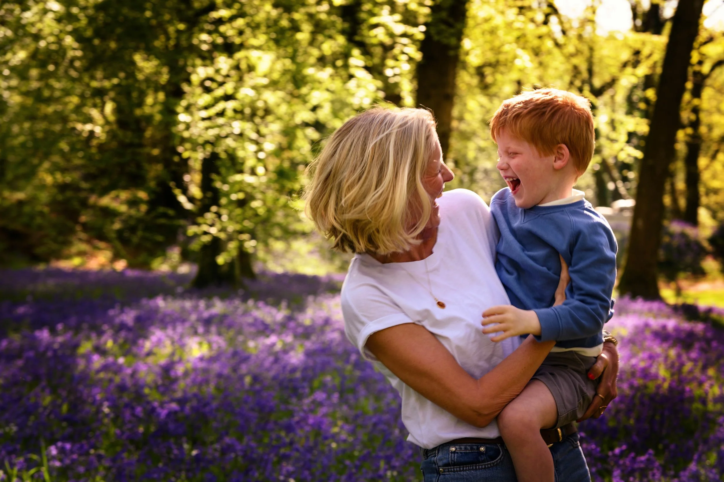 Mother holds her young son in her arms as they both share a joke in a bluebell wood in Devon