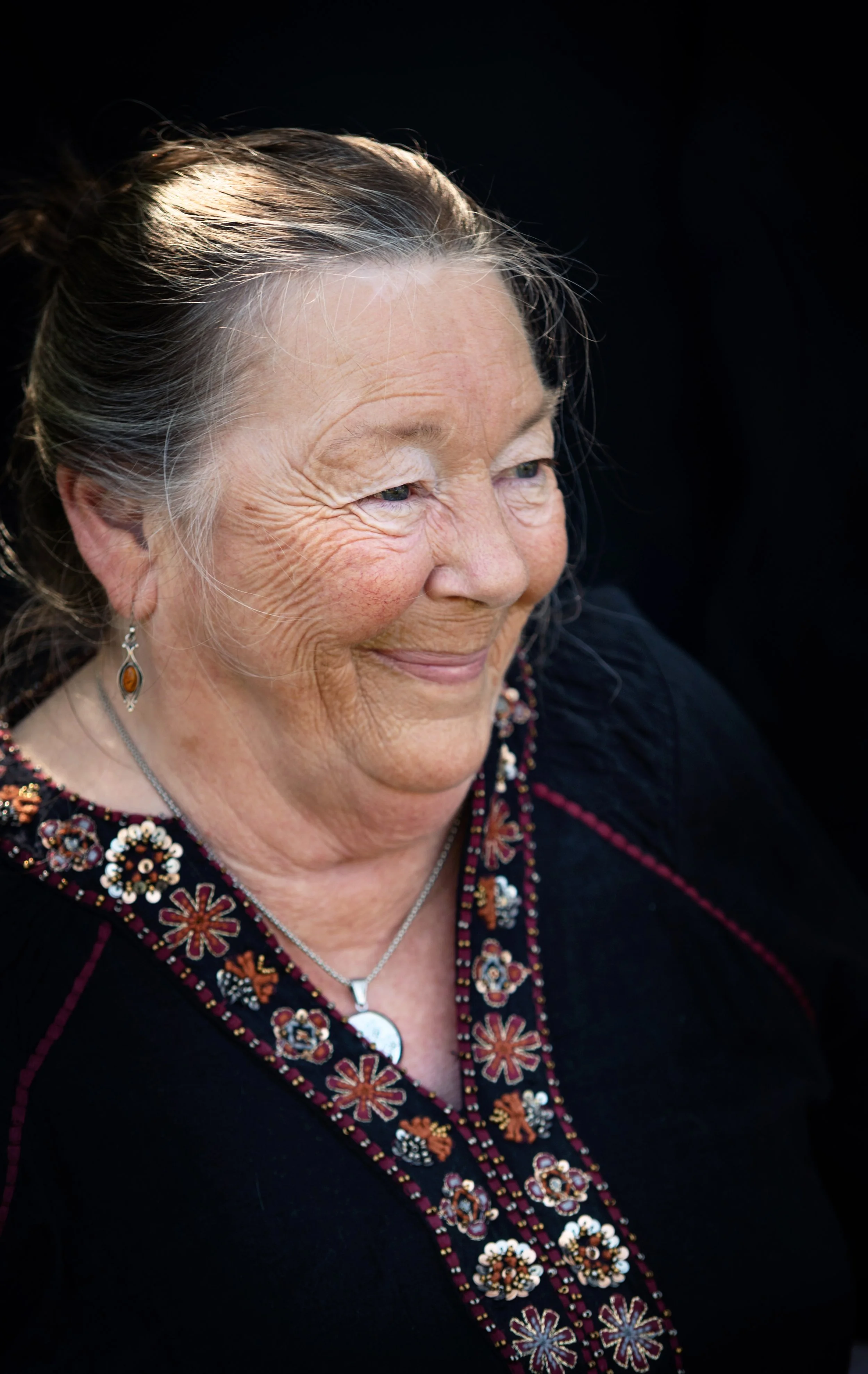 Close-up of an elderly woman with gray hair pulled back, smiling gently, wearing jewelry and a black outfit with colorful embroidery.