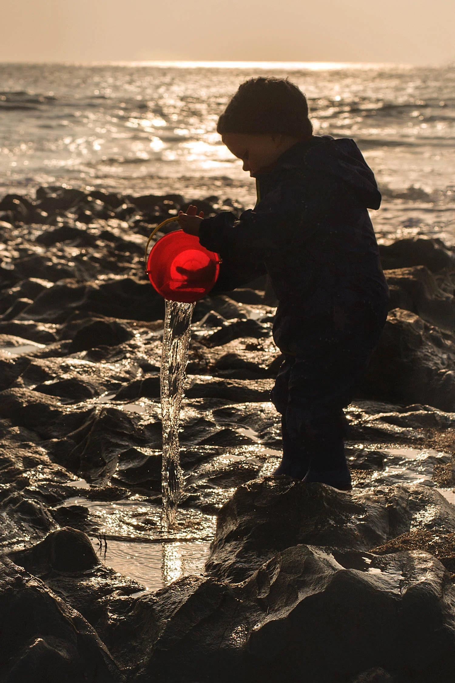 little boy pours sea water out of sunlit red bucket