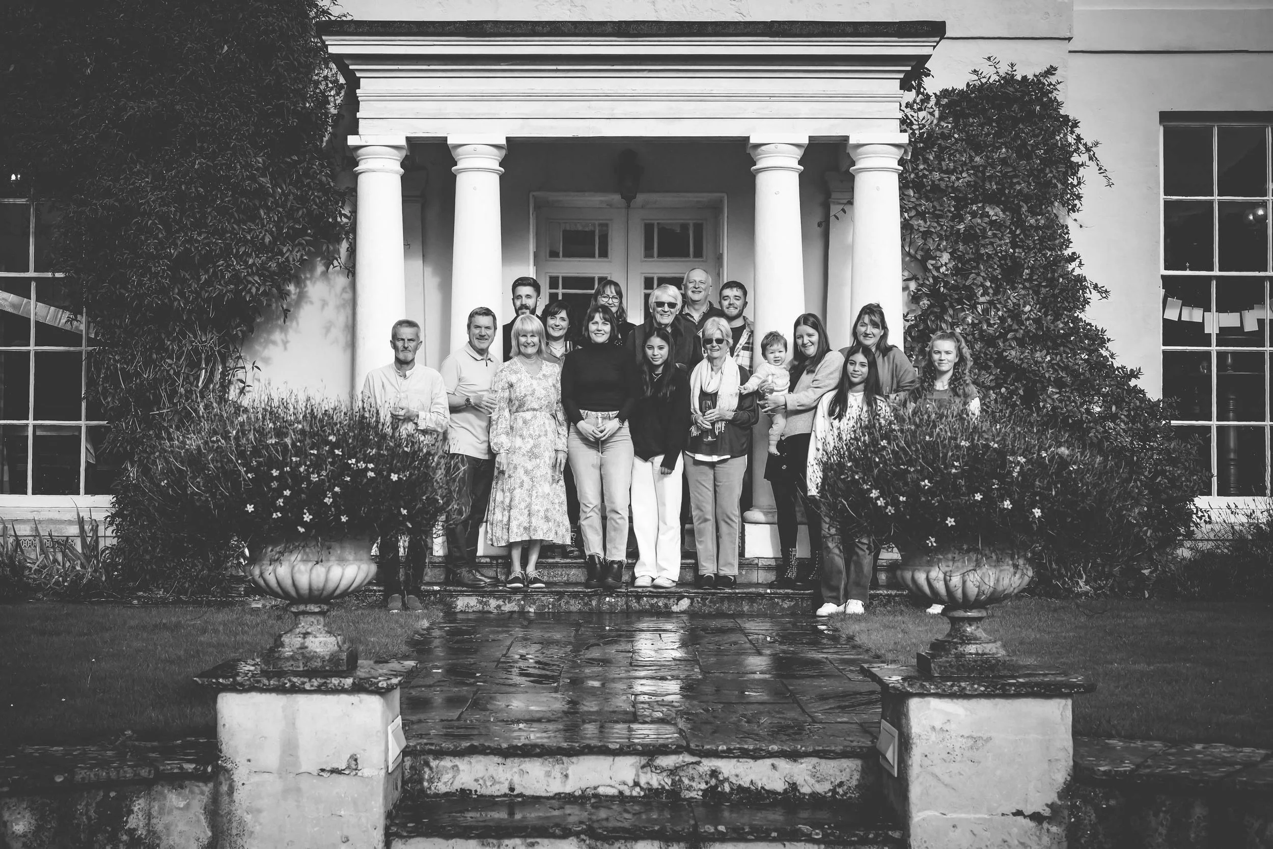 Black and white photo of a large family group standing on front steps of a house with white columns, surrounded by large potted plants and bushes.