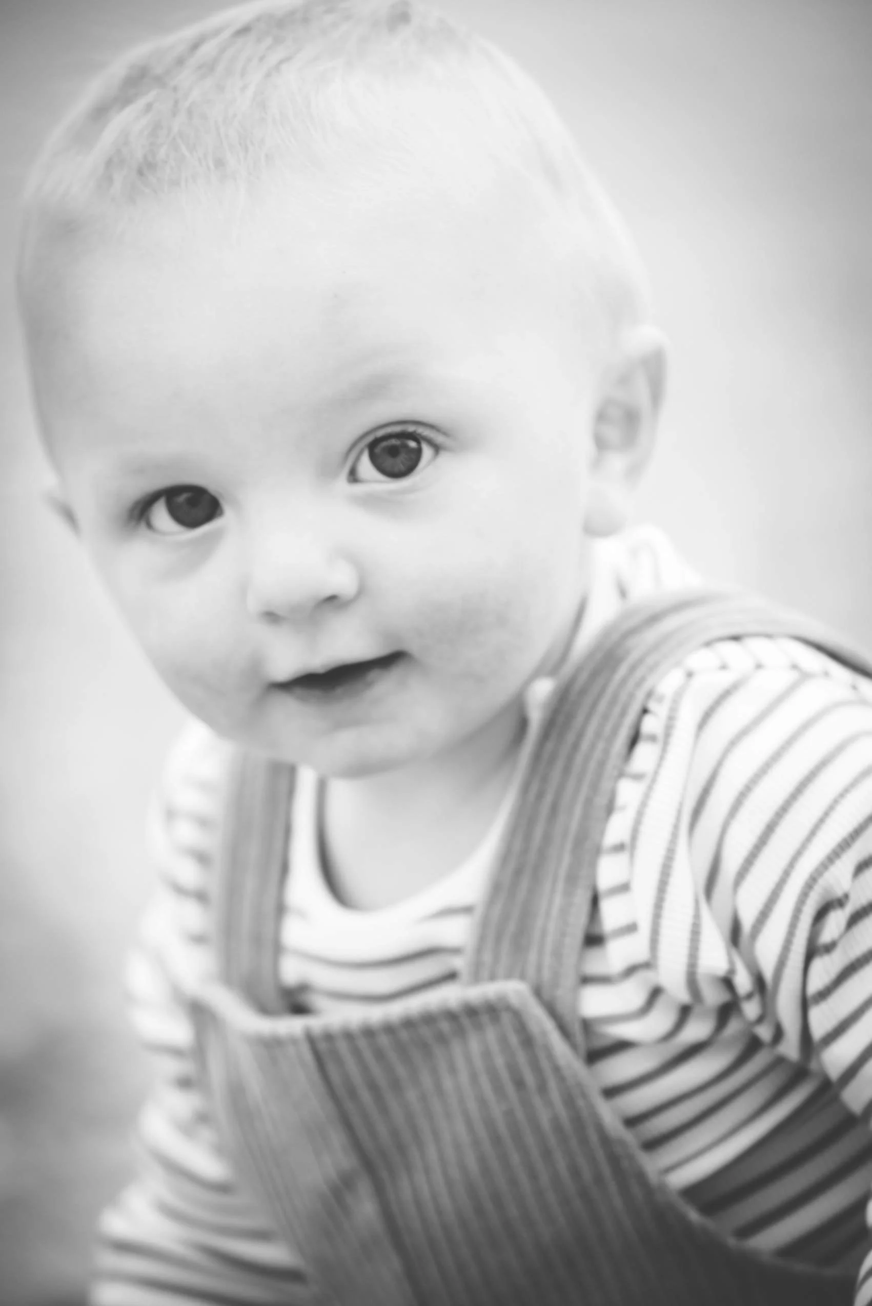 Black and white photo of a young child with light hair, wearing a striped shirt and overalls, looking at the camera with a curious expression.