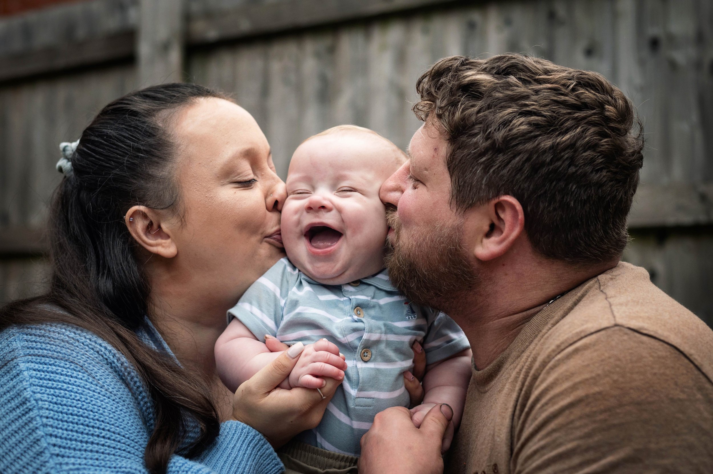 A woman and a man kissing a smiling baby's cheeks in an outdoor setting with a wooden fence background.