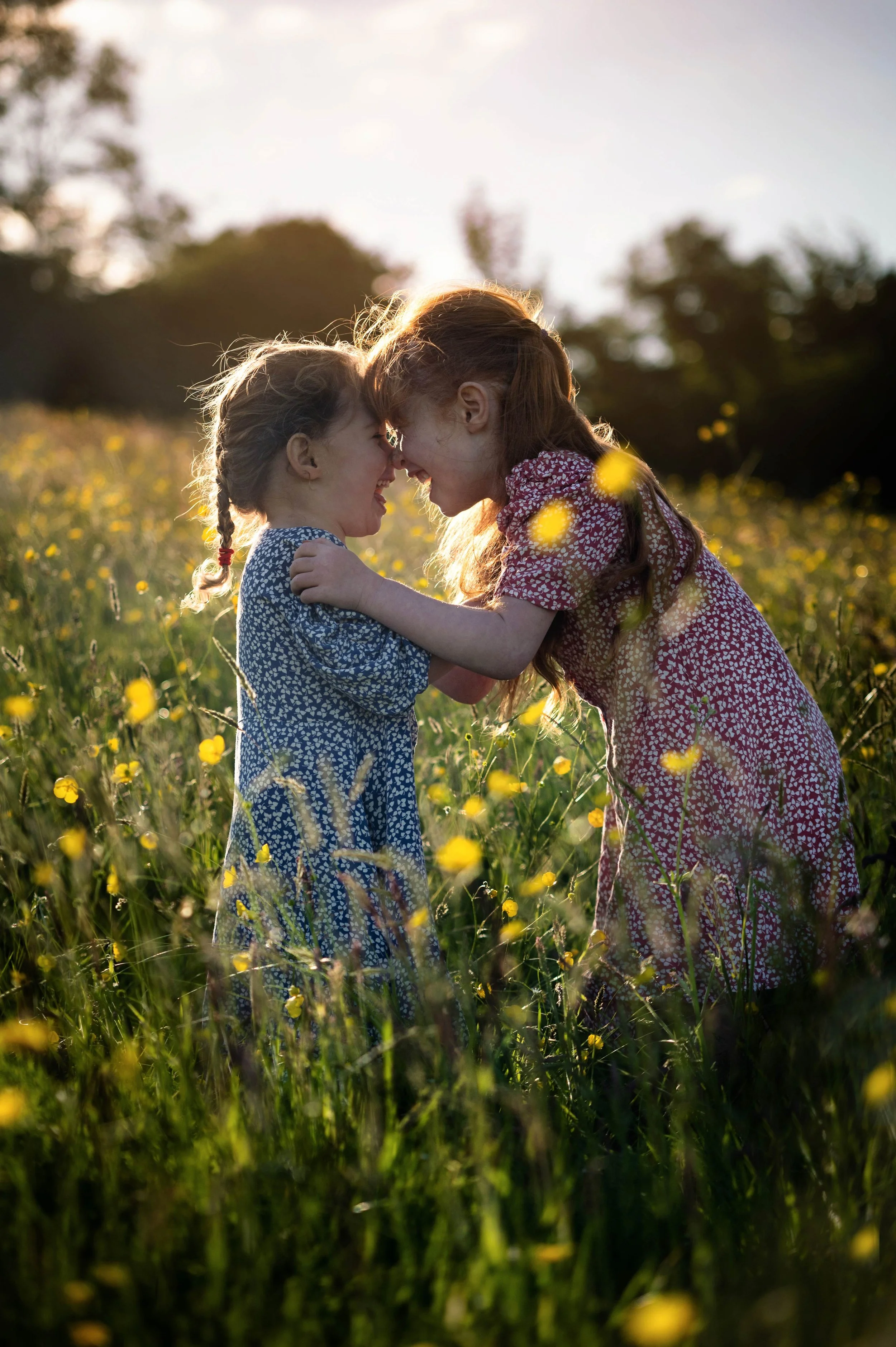 Two young girls, one with braided hair and the other with loose hair, smiling and touching foreheads in a field of yellow flowers during sunset.
