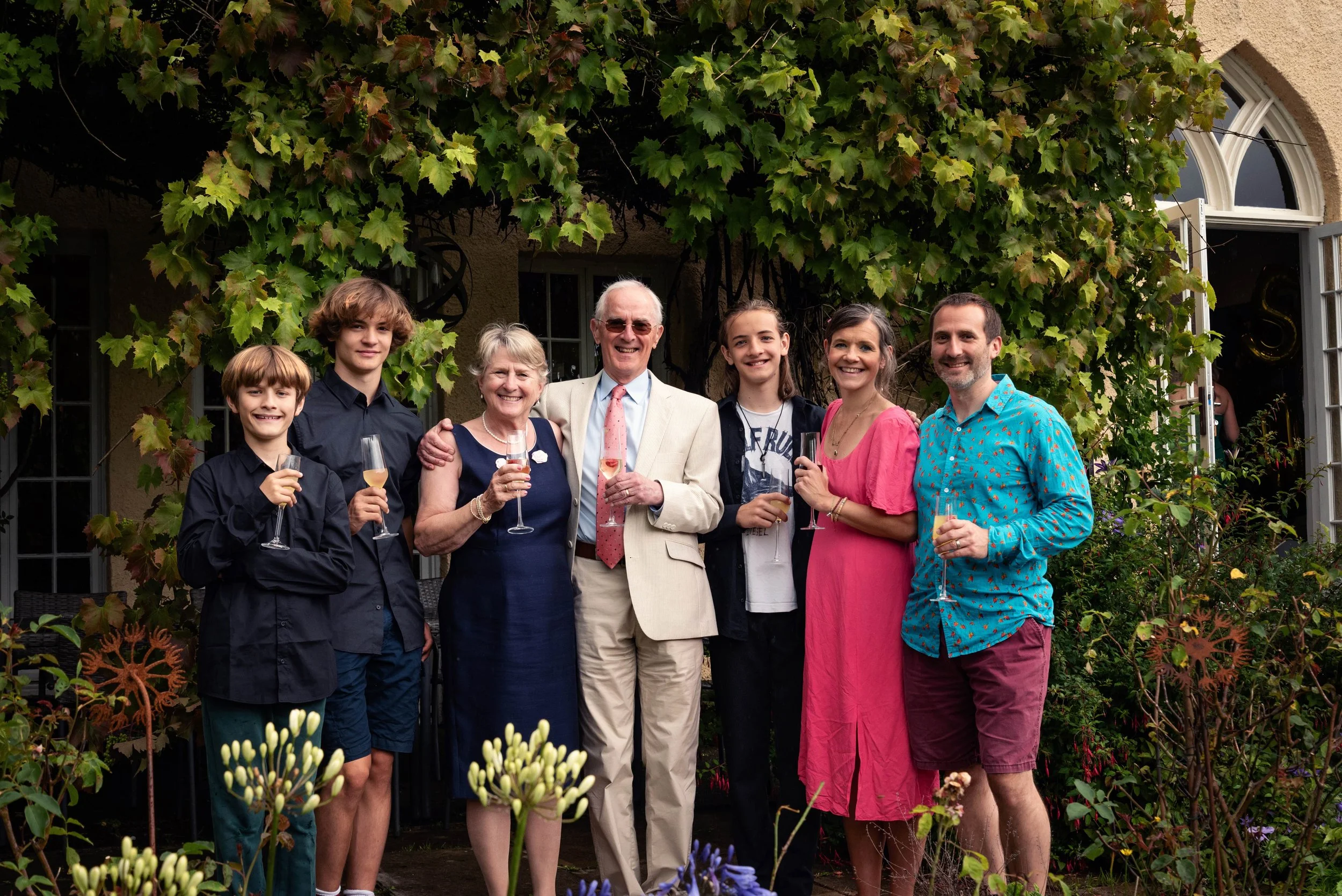 Family and friends gathered outdoors at a celebration, standing together for a group photo, holding champagne glasses, under green leafy vines in a garden setting.
