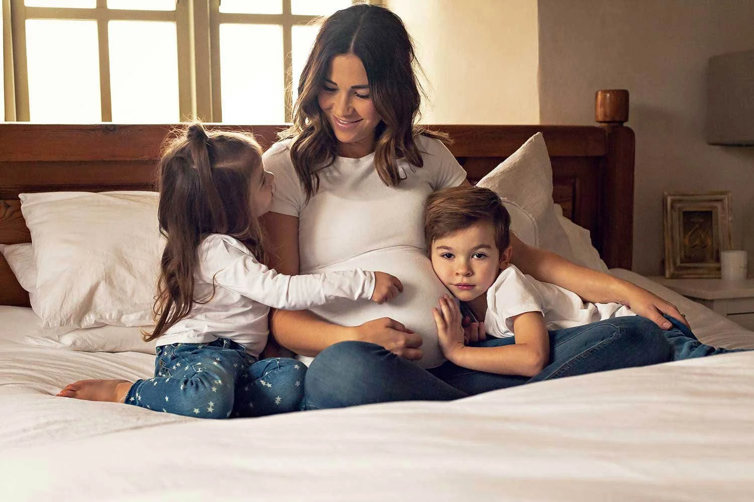 A pregnant woman sitting on a bed with her two young children, a girl and a boy, in a warmly lit bedroom. The girl is touching the woman's pregnant belly and the boy is resting his head on her lap, both children are looking at her.