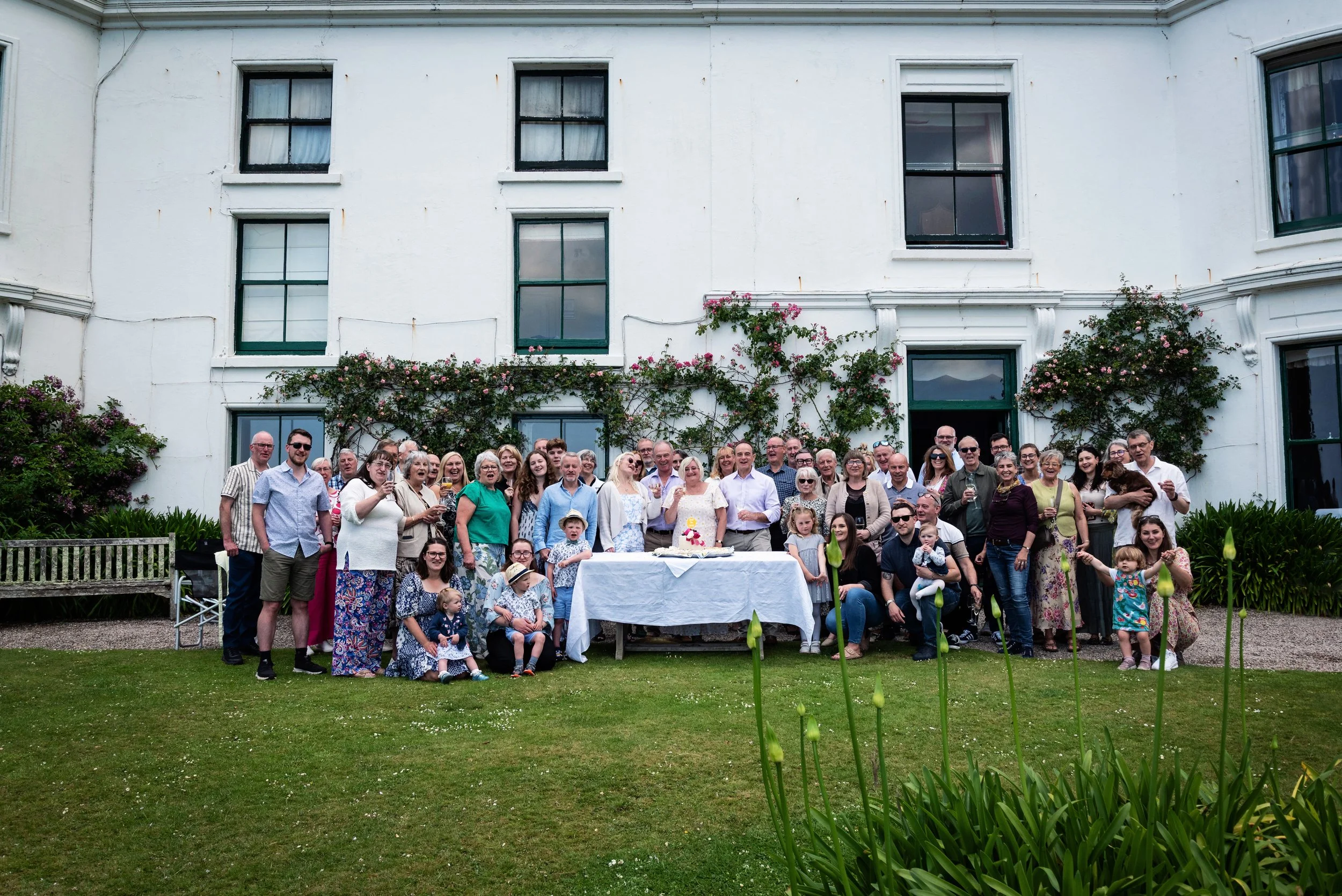 A large group of people standing outdoors in front of a white building with greenery and flowers, gathered around a table with a cake, celebrating a special occasion.