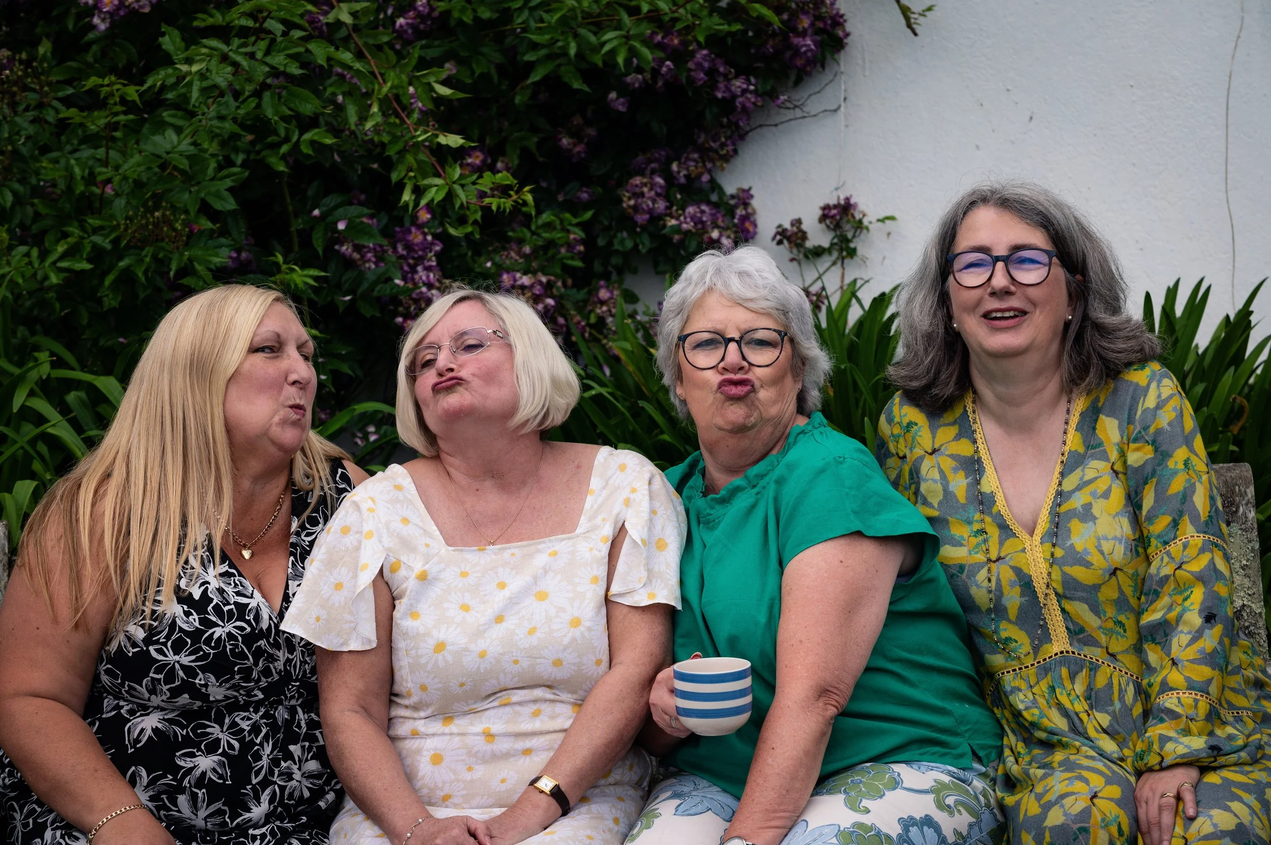 Four women sitting outdoors in front of green leafy bushes and purple flowers, making funny faces at the camera.