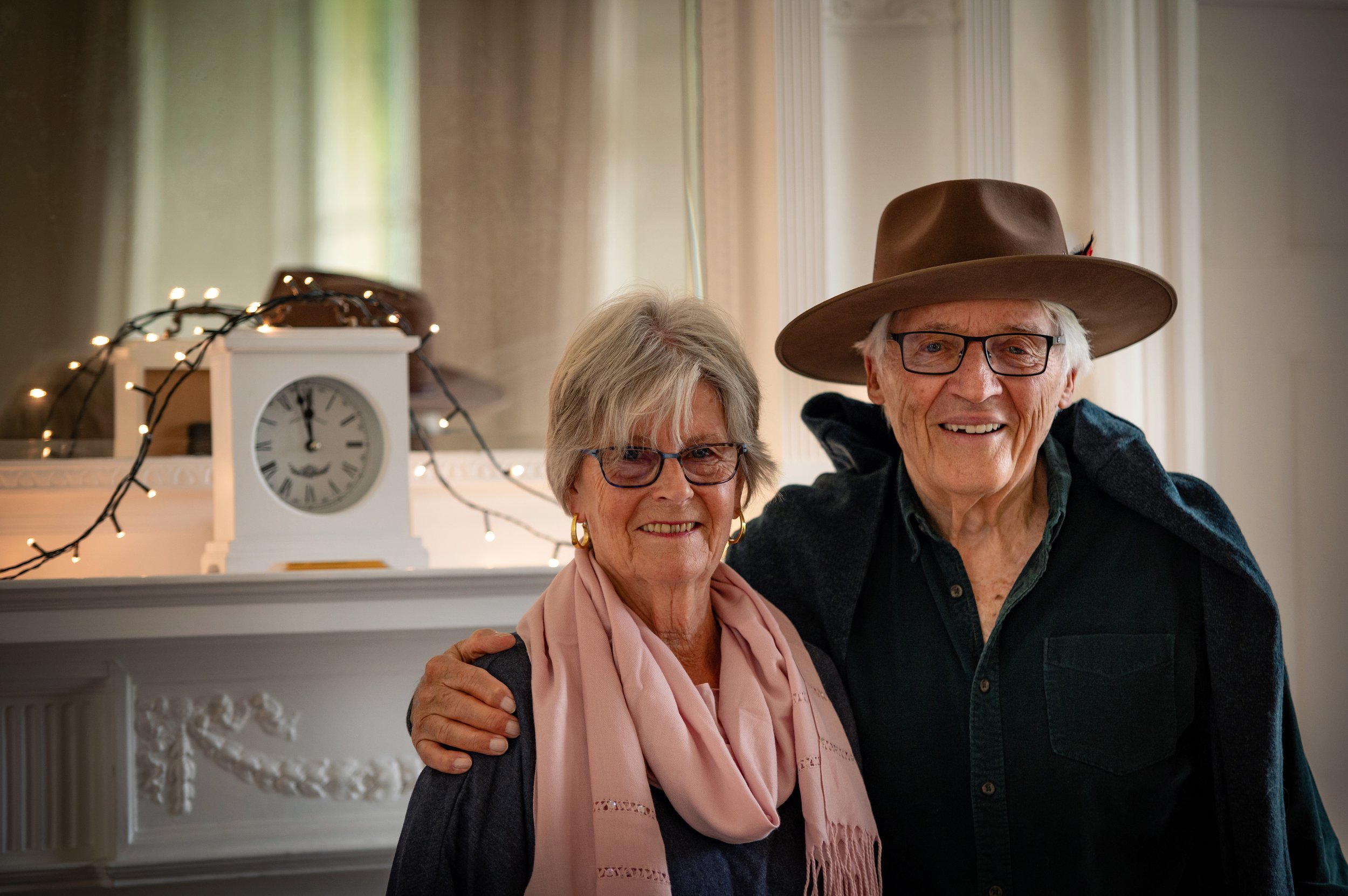 An elderly couple smiling indoors, with the woman wearing glasses, gold earrings, a pink scarf, and a dark sweater, and the man wearing glasses, a brown hat, and a dark shirt. They are standing close together in front of a white mantle with a clock a