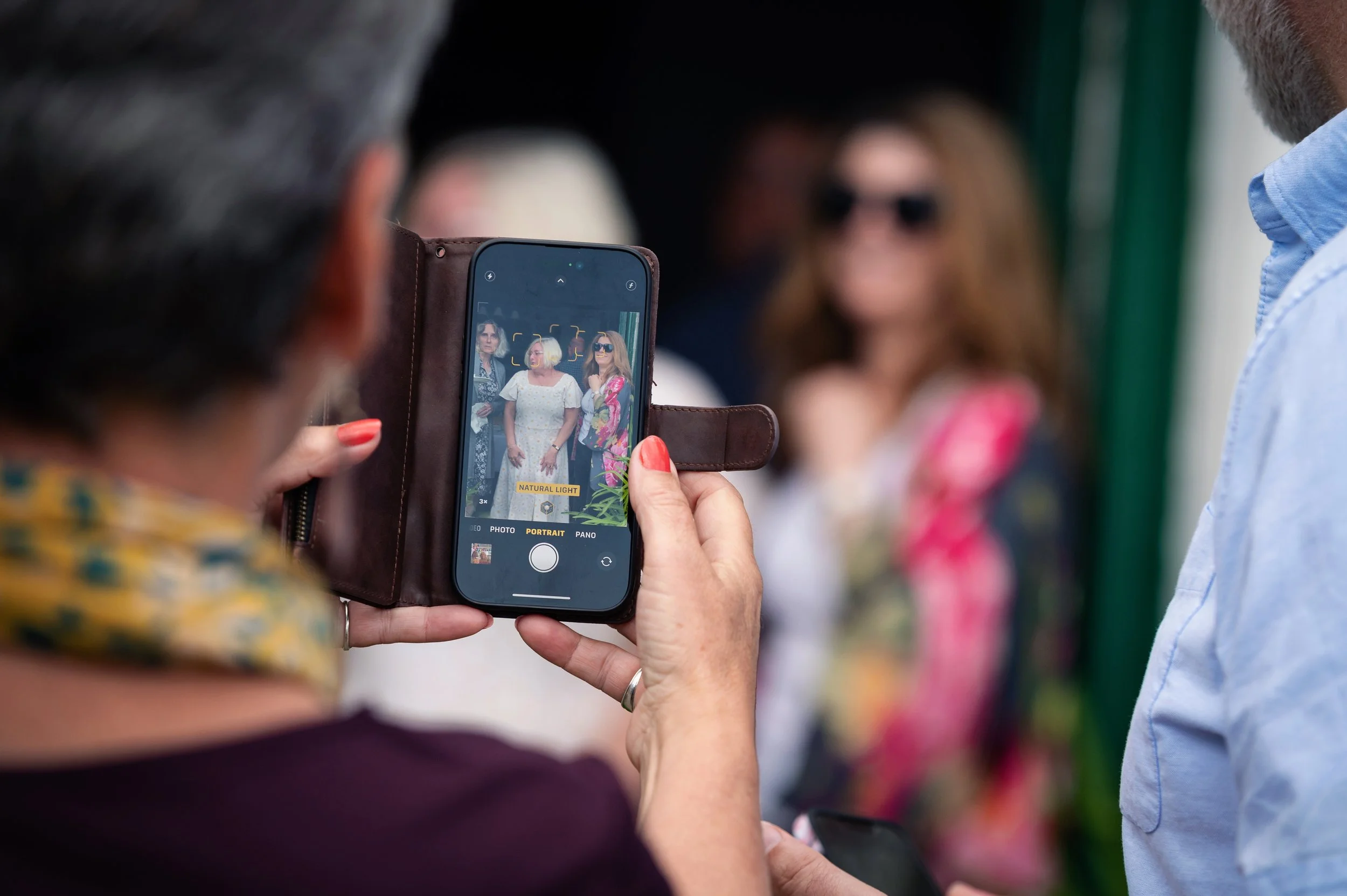 A woman taking a photo of three women standing against a green and black background with her cellphone.