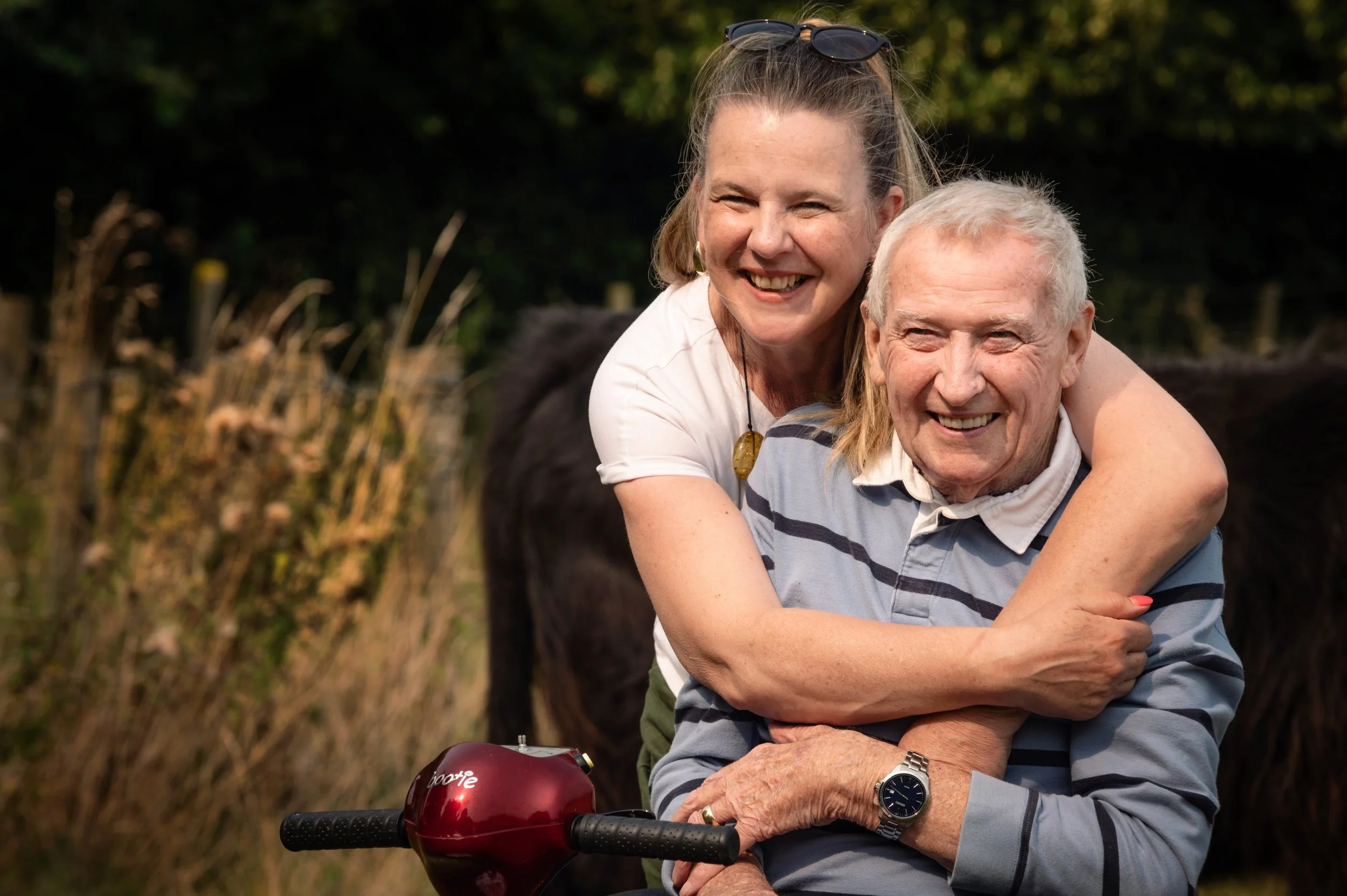 A smiling elderly man is sitting on a mobility scooter with a woman hugging him from behind. They are outdoors in a natural setting, with a fence and trees in the background.