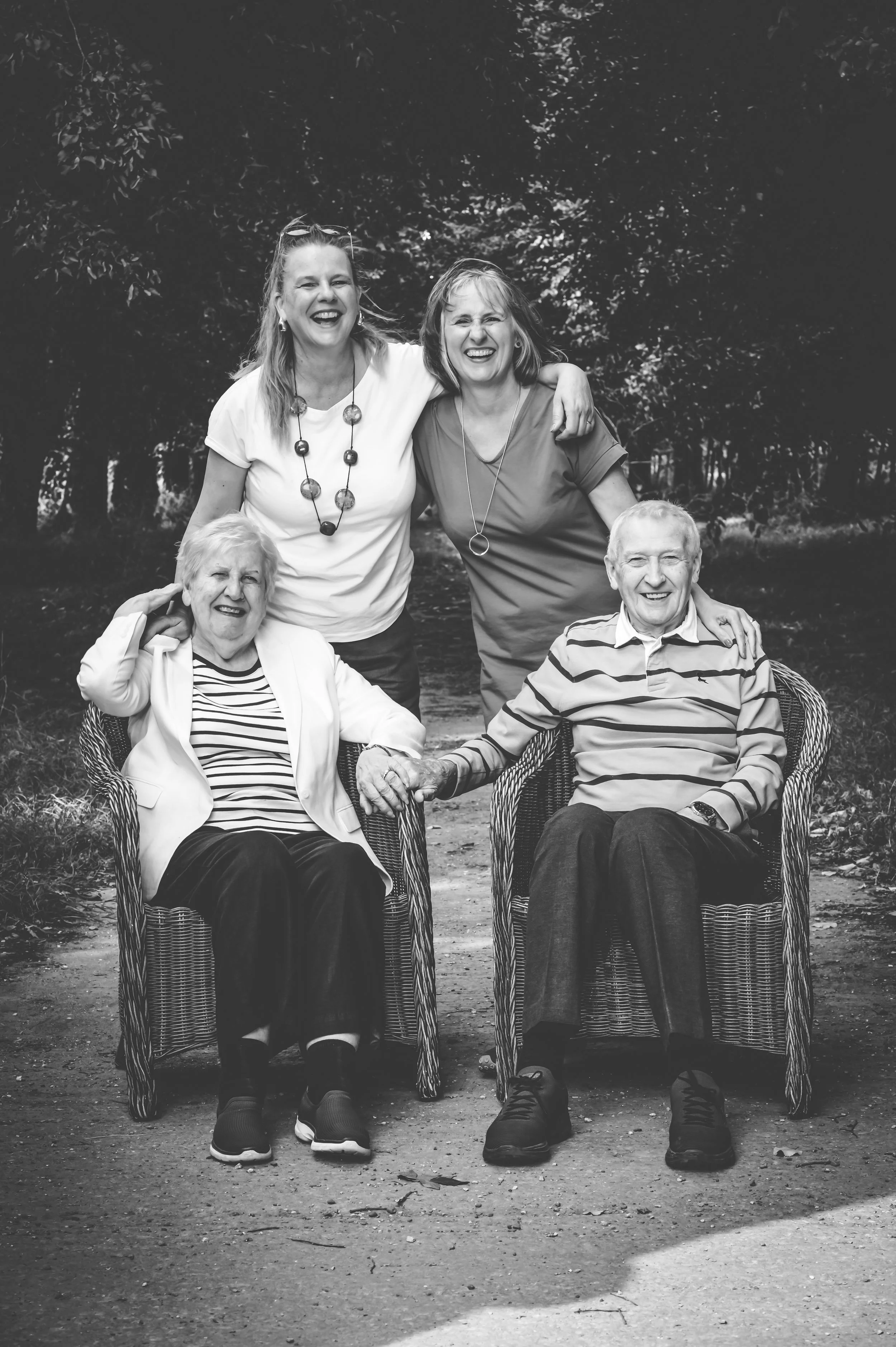 Black and white photo of four women, two elderly sitting in wicker chairs, two middle-aged standing behind, outdoors in a wooded area, smiling and holding hands.