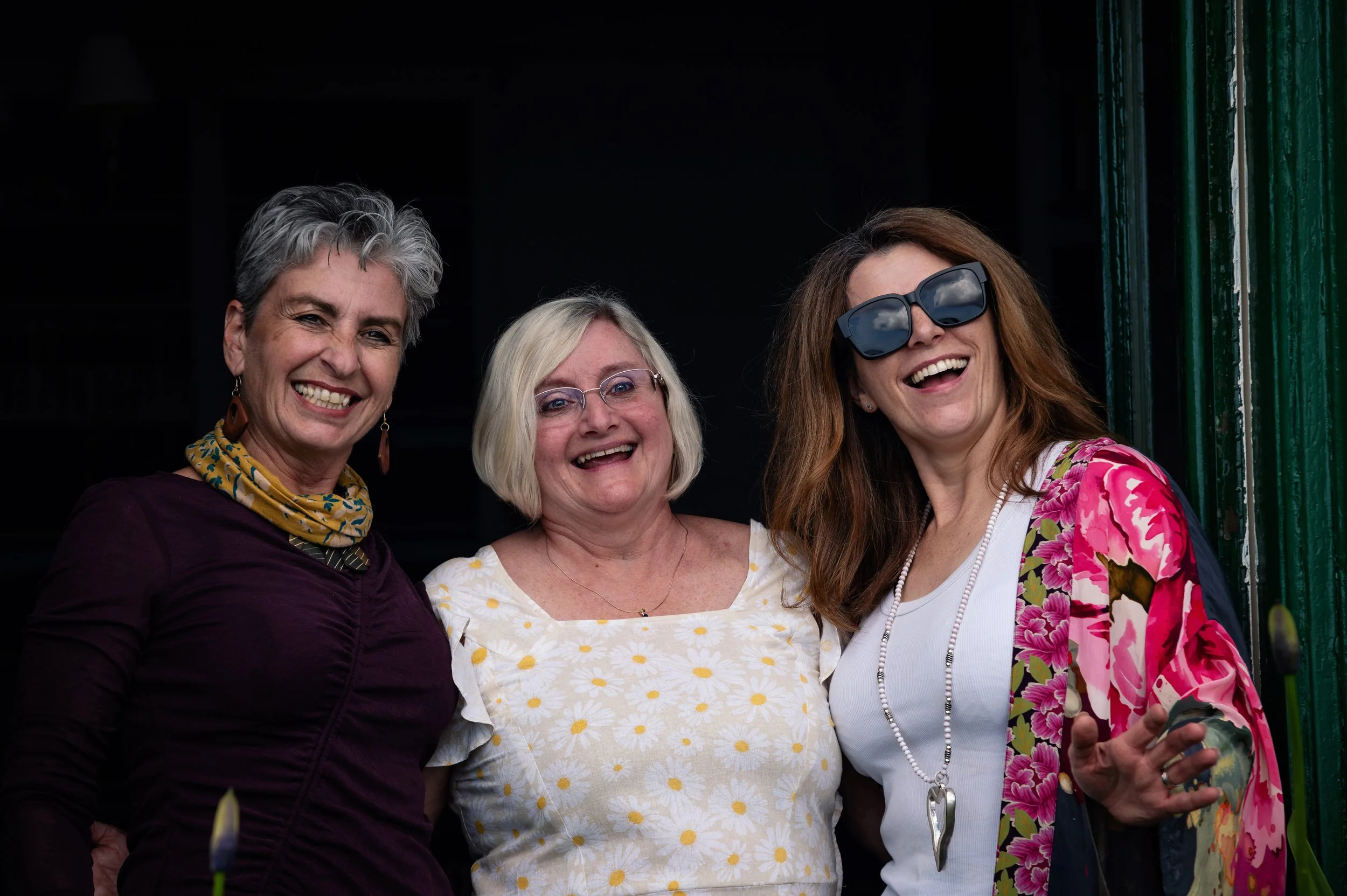 Three women smiling and standing close together, dressed casually, with dark background and some greenery visible.