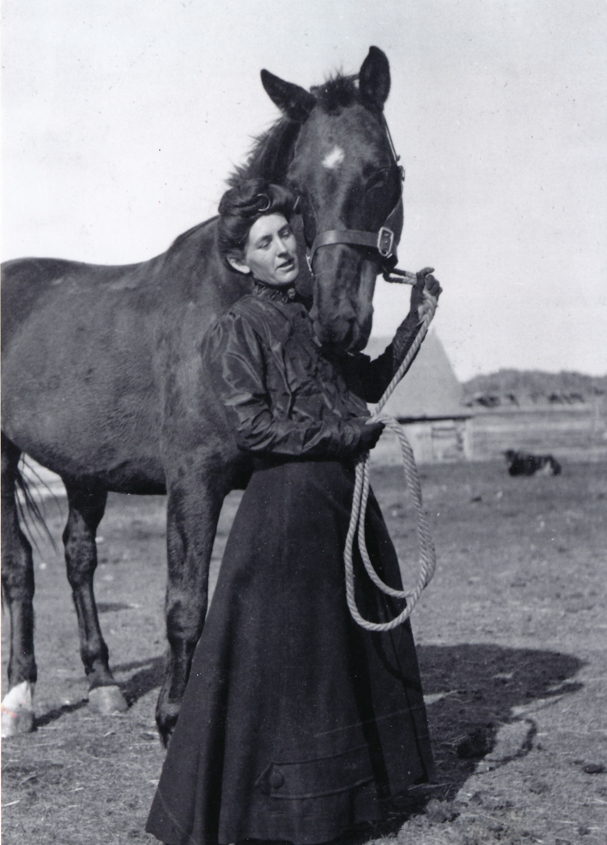 From the Glenbow Archives (Image: A young woman holding a loop of halter rope while being nuzzled by her black horse which has a white blaze between its eyes. She is wearing a long black riding skirt, dark high-necked blouse and black gloves. In the background is a barn and corral.)