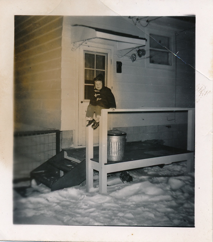 1955 Bruce aged three on the back porch of the new house at 7403 - 21A St. S.E. in Ogden on the outskirts of Calgary. (Image: A young boy in a dark-coloured parka sitting somewhat precariously on a porch railing of a small house in the background with snow covering the foreground.)
