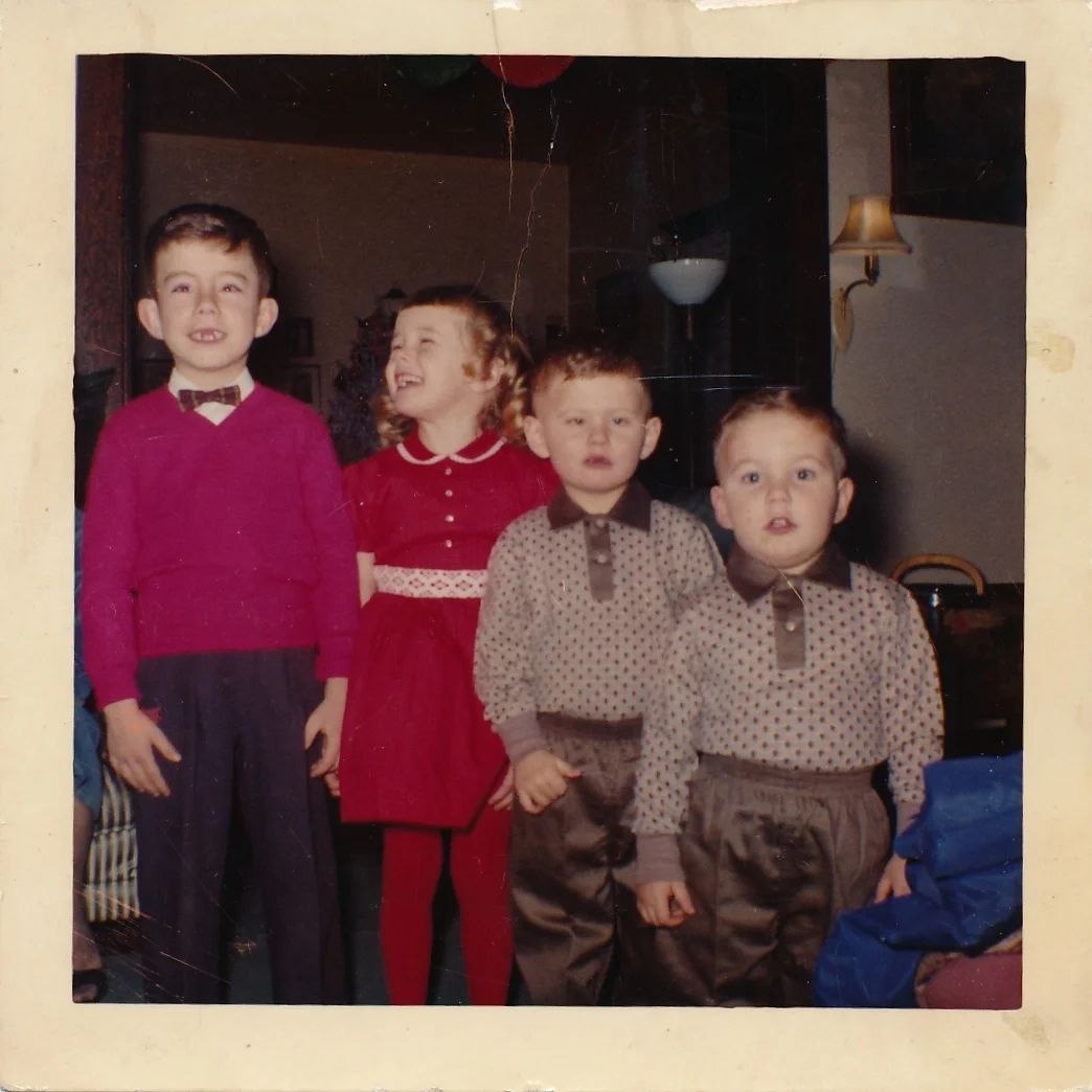 1960 Bruce aged six in a bow tie and red sweater, sister Susan aged five in a red dress, brothers Michael aged four and Allan aged three. (Image; Four children, three boys and one girl all dressed in Christmas garb and the two youngest boys are wearing matching outfits of patterned shirts and shiny pants. The girl is smiling up at her older brother who is missing two front teeth.)